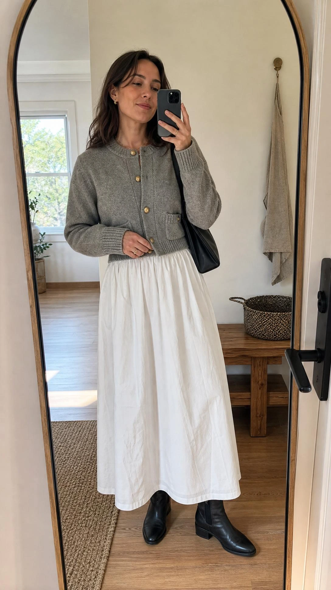 Mirror selfie of a woman styling white maxi skirt outfit ideas with a cardigan jacket and ankle boots in a clean entryway