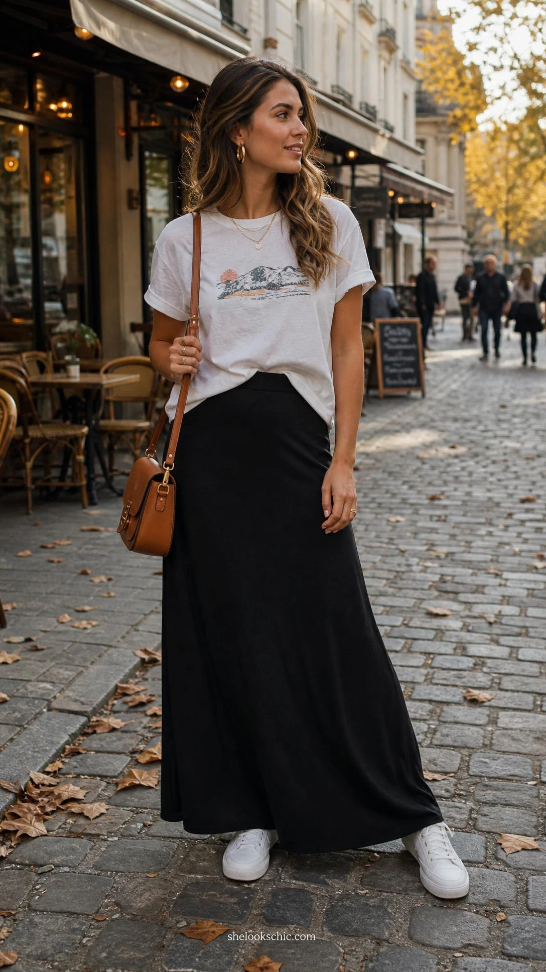 Woman half-tucking a faded graphic tee into a flowing black maxi skirt on a sun-lit cobblestone street, effortless casual style
