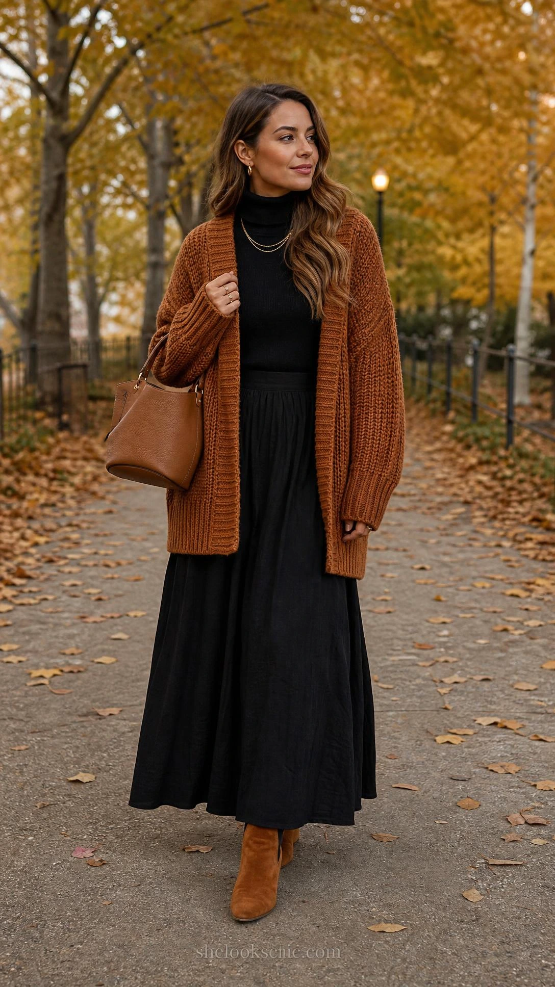 Woman in flowing black maxi skirt and cozy cardigan with ankle boots walking autumn leaf-scattered park path among golden trees