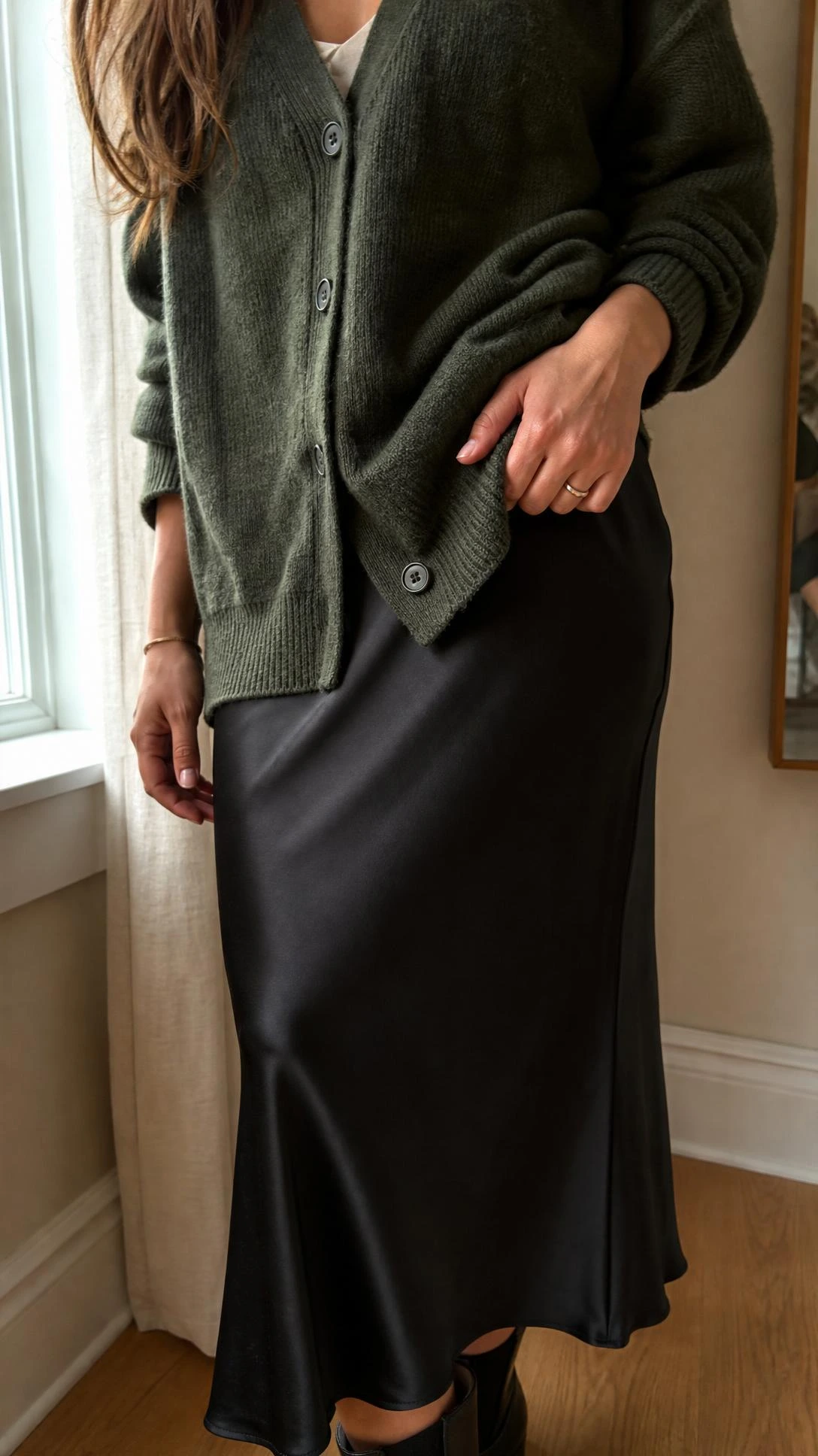 Close-up of a woman by a bright window wearing a forest green cardigan with a black slip skirt, highlighting rich knit texture and sleek contrast.