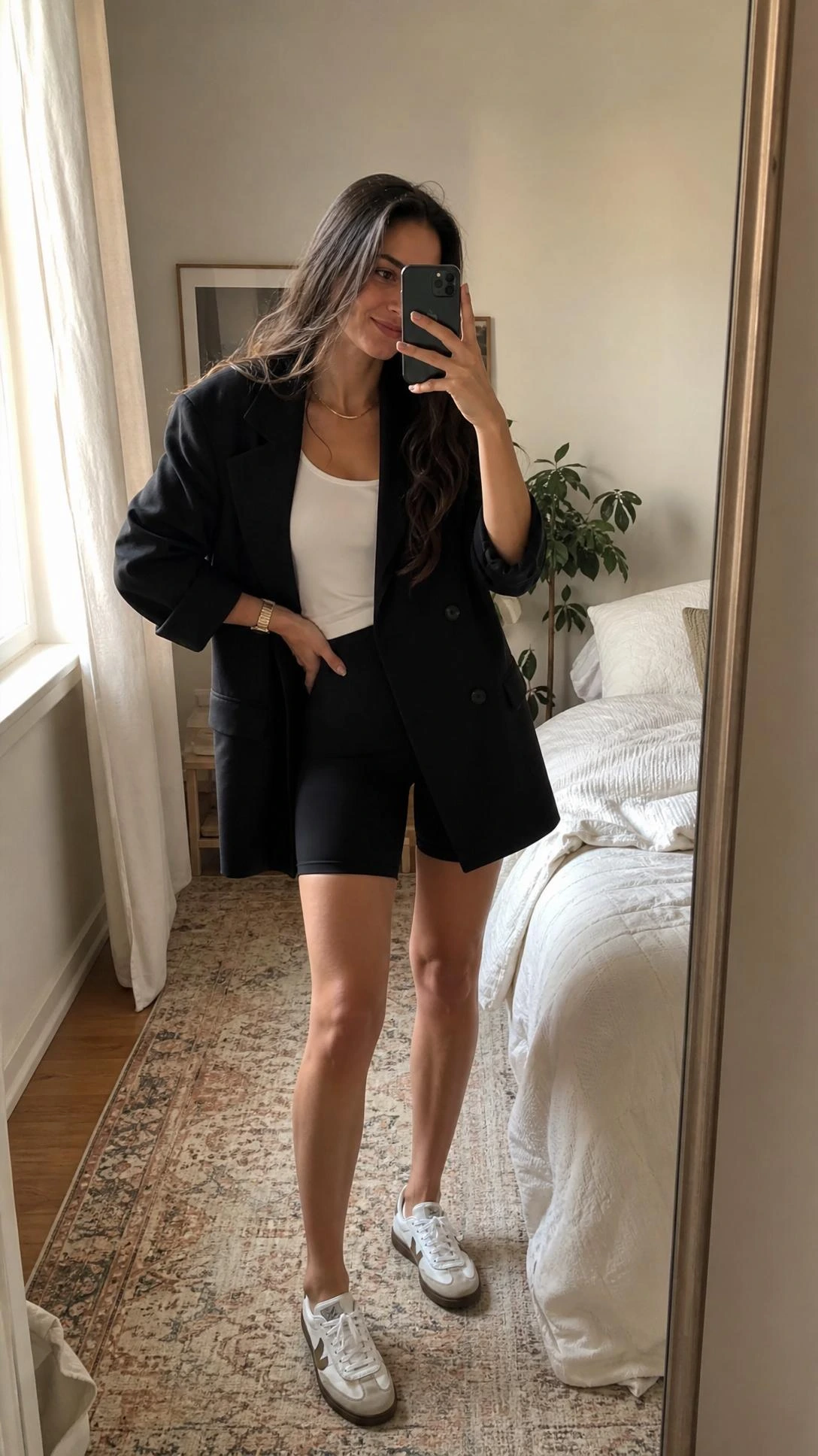 Mirror selfie of a tall slim woman in an oversized blazer, biker shorts, and white tank in a softly lit bedroom.