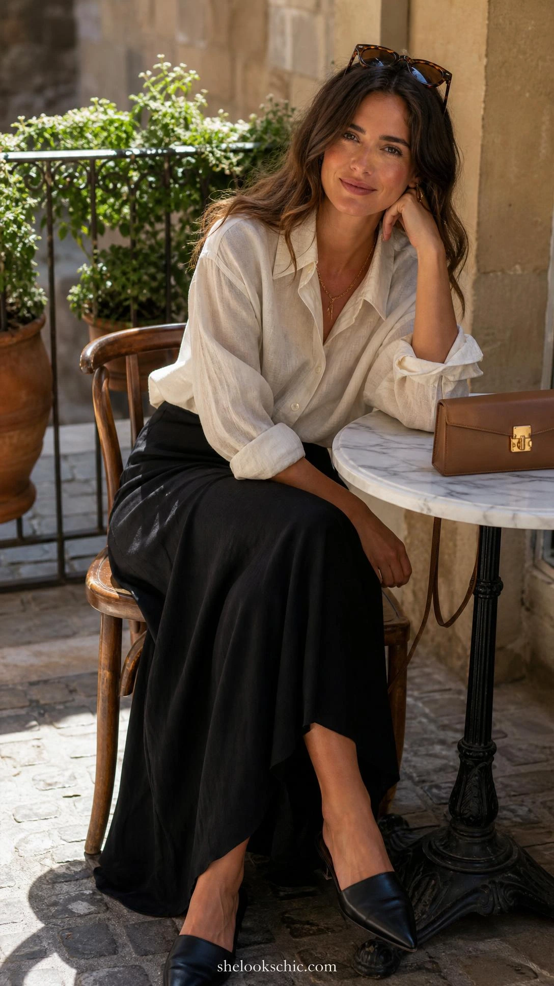 Woman in oversized ivory linen button-down shirt tucked into a black maxi skirt, seated at a sunlit European café terrace showing effortless casual chic