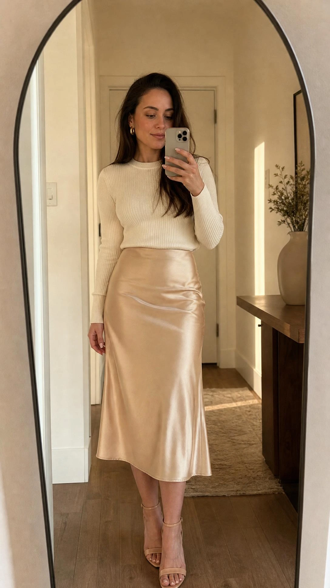 Woman styling a satin midi skirt with a fitted ribbed knit top for a cocktail outfit, captured in a mirror selfie in a softly lit home entryway