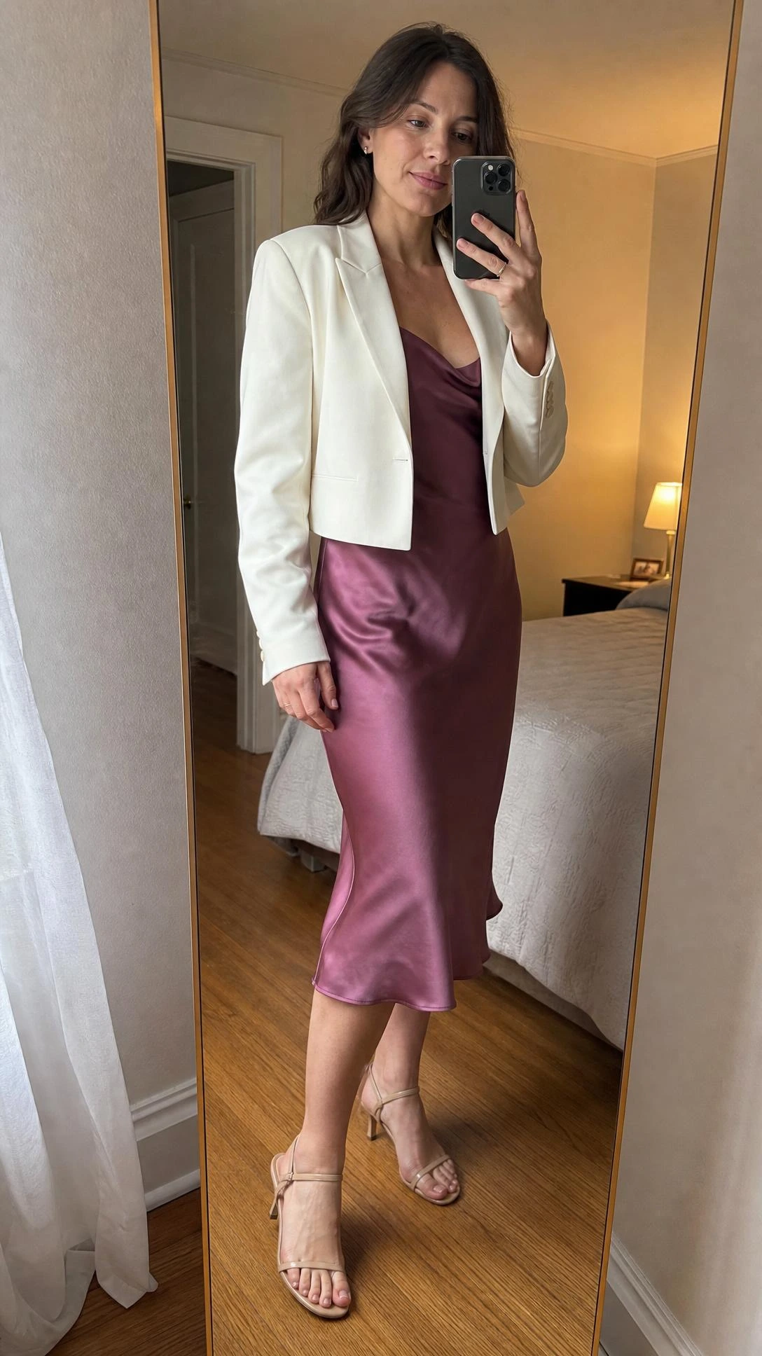 Woman in slip dress with cropped structured jacket poses for mirror selfie in warmly lit bedroom, showing a polished cocktail outfit combination