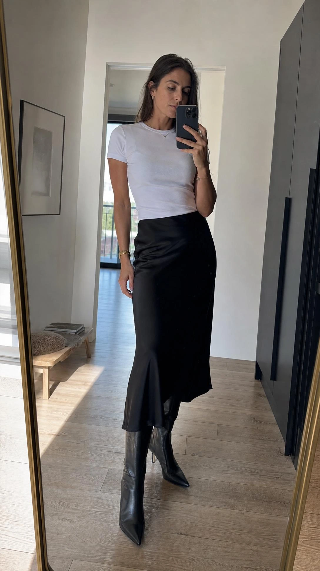 Angled mirror selfie of a tall slim woman in a slip midi skirt, fitted tee, and tall boots in natural daylight.
