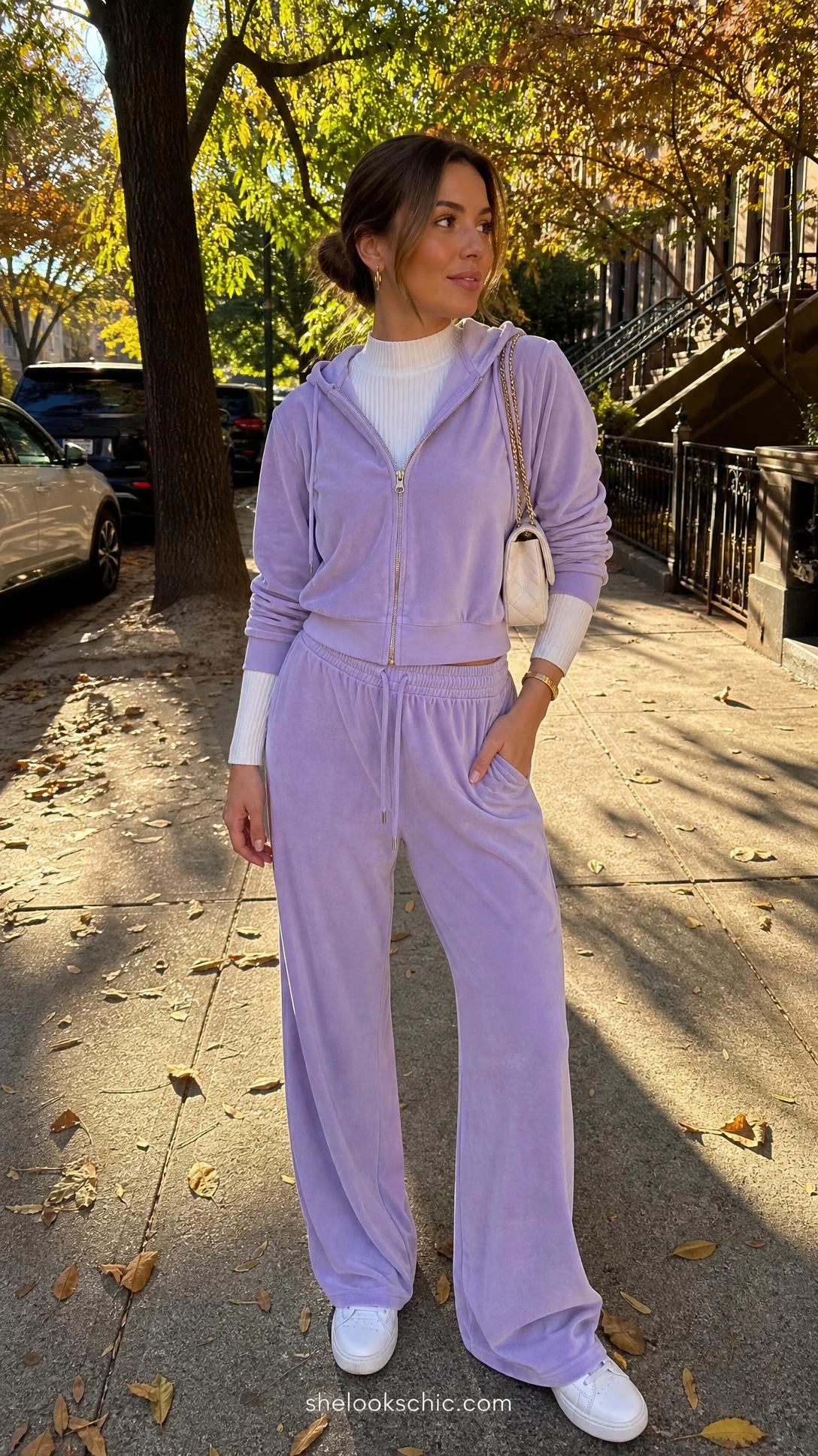 Woman wearing a lavender velour tracksuit on a tree-lined sidewalk, capturing the iconic Y2K outfit trend in a soft pastel hue