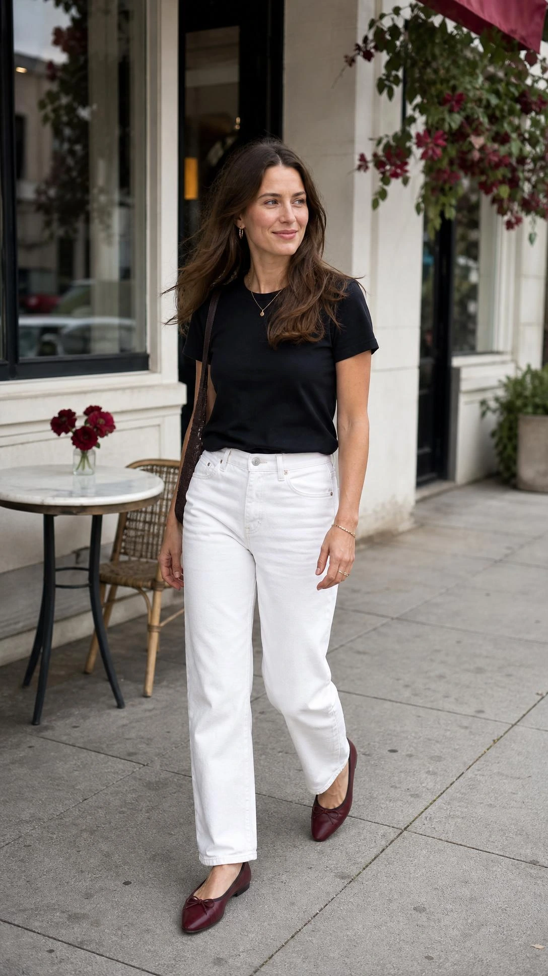 Woman walking outside a cafe in one of the white jeans outfit ideas: a black tucked tee and burgundy flats