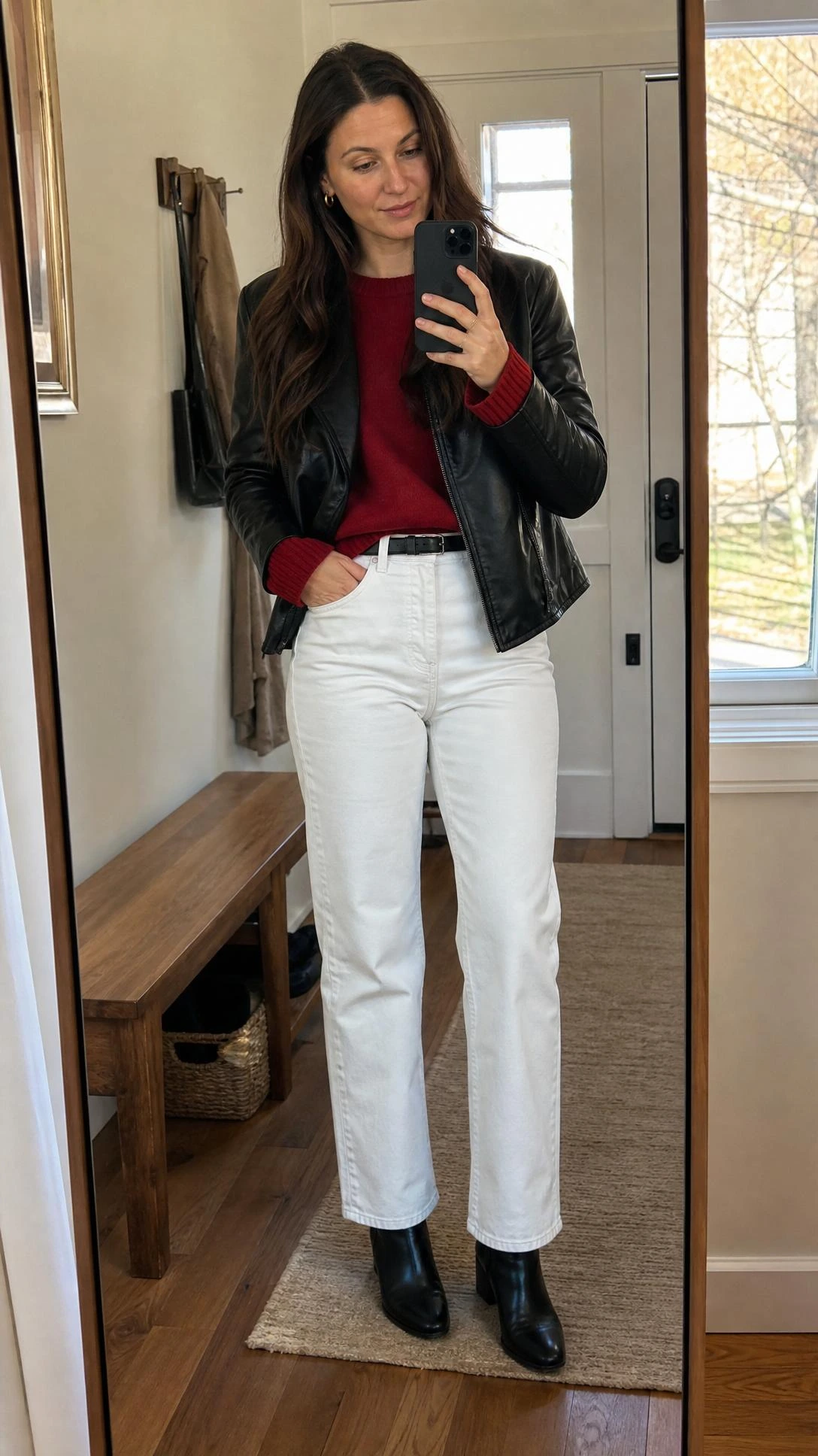 Woman takes an entryway mirror selfie in a red sweater, black leather jacket, and white jeans outfit ideas look