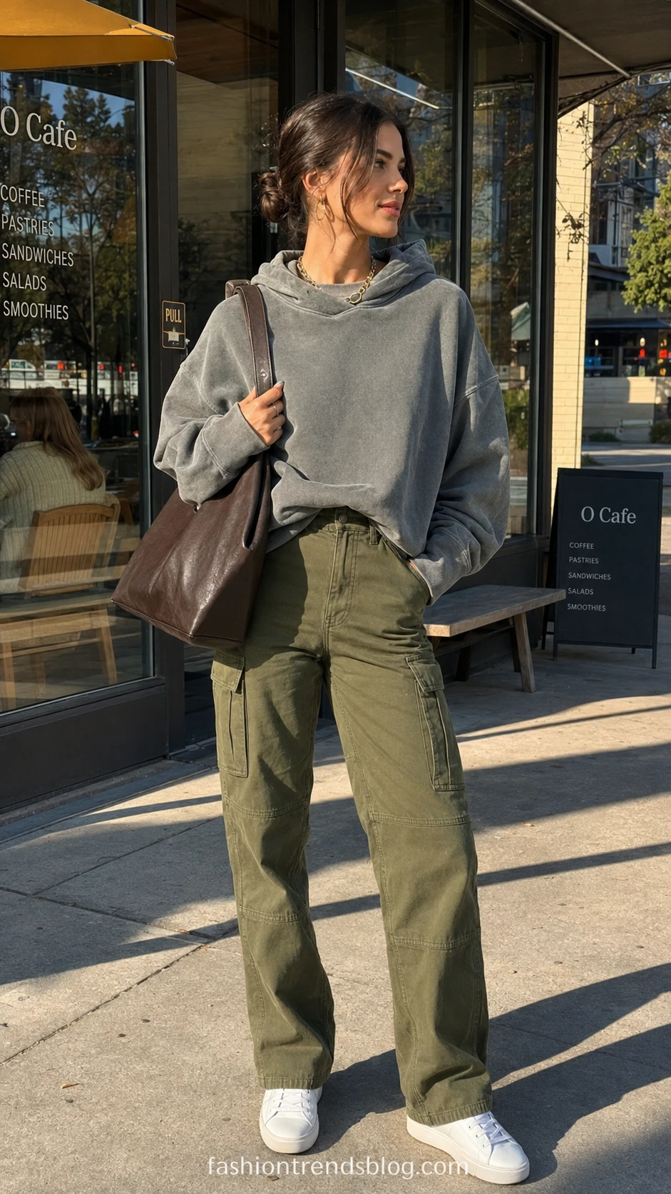 Woman wearing olive green cargo pants with a white sports bra and oversized hoodie on a sunny urban sidewalk, showing effortless athleisure street style