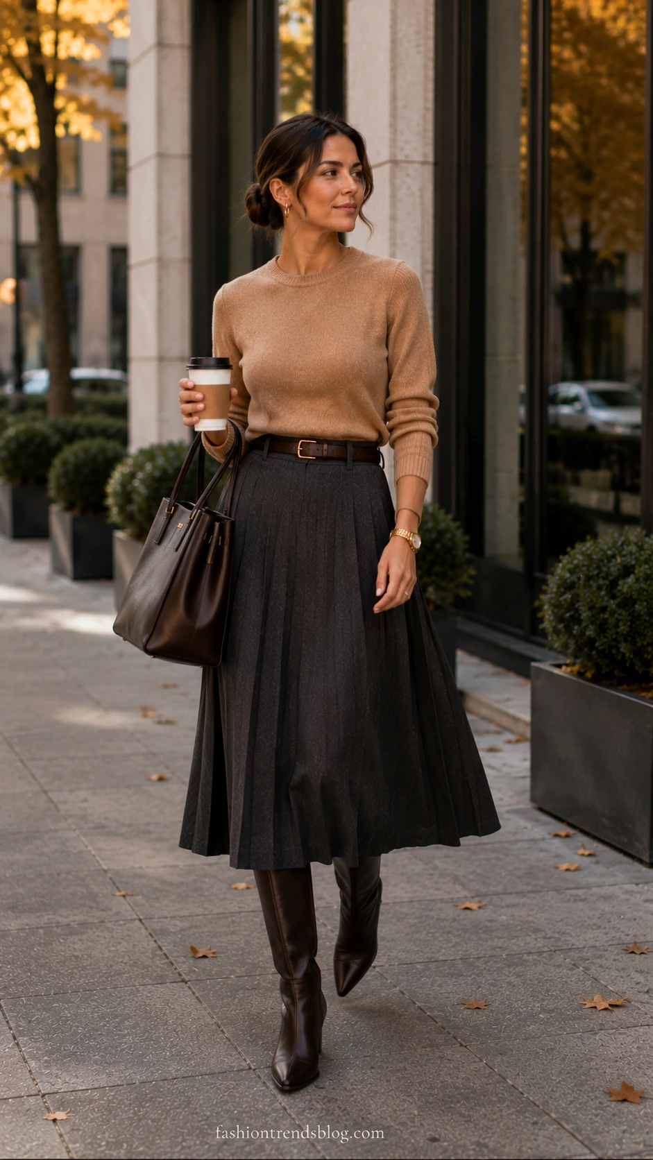 Woman in a fall office work outfit: camel sweater tucked into a charcoal pleated midi skirt with brown boots.