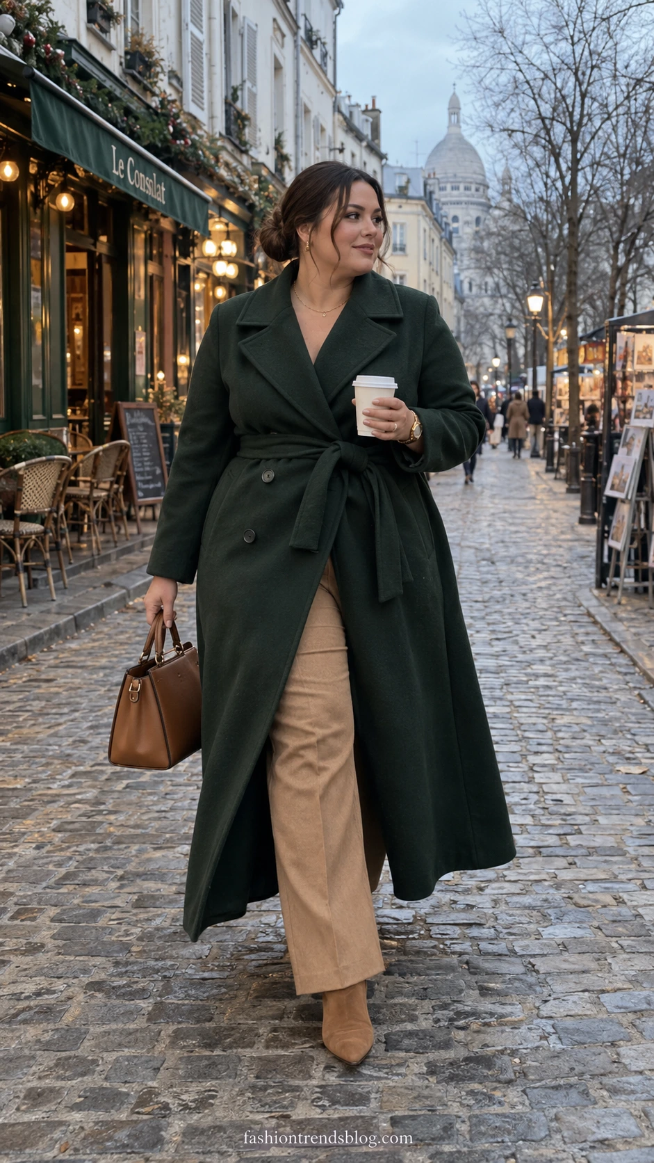 Plus size woman in a belted wrap coat and tailored trousers walking a cobblestone street with glowing cafe lights and bare winter trees