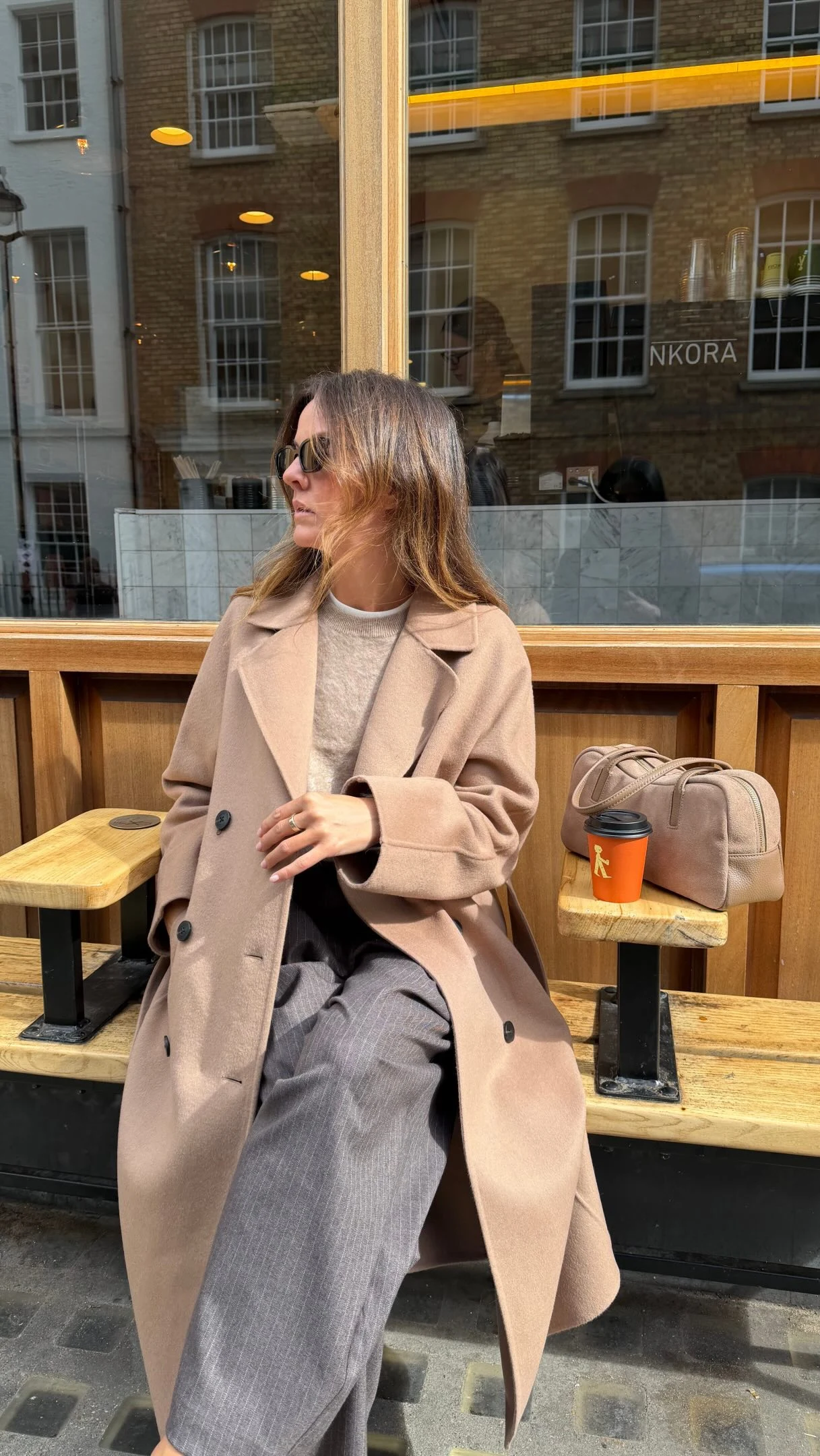 Seated woman in pale beige coat, wide-leg gray trousers, and dark sunglasses with beige handbag.