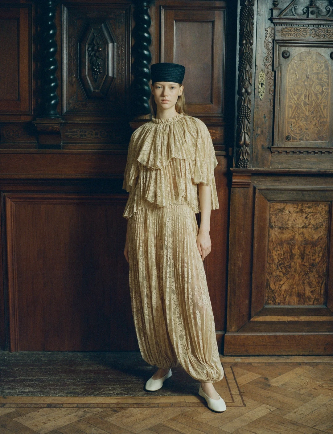 Person wearing a beige ruffled lace dress with a dark flat-topped hat and white shoes.