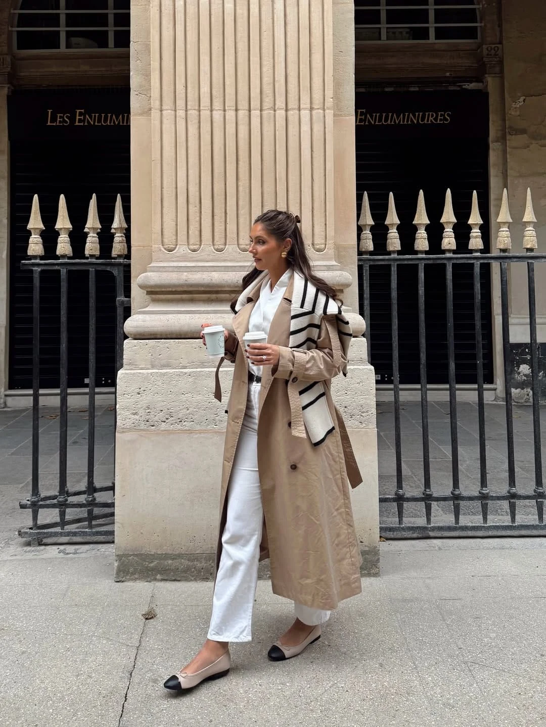 Beige trench coat layered over white shirt and cropped pants with a striped knit draped over the shoulders.