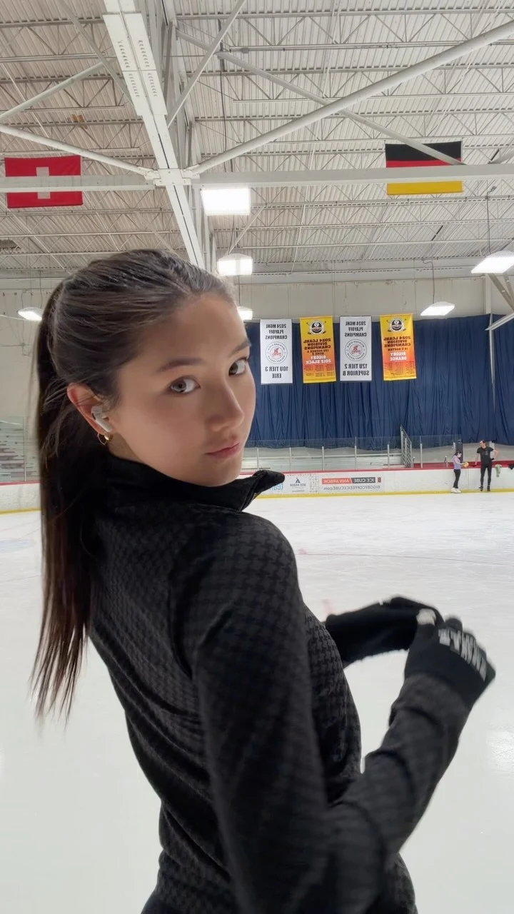 Person in a fitted black checkered jacket and black gloves at an indoor ice rink.