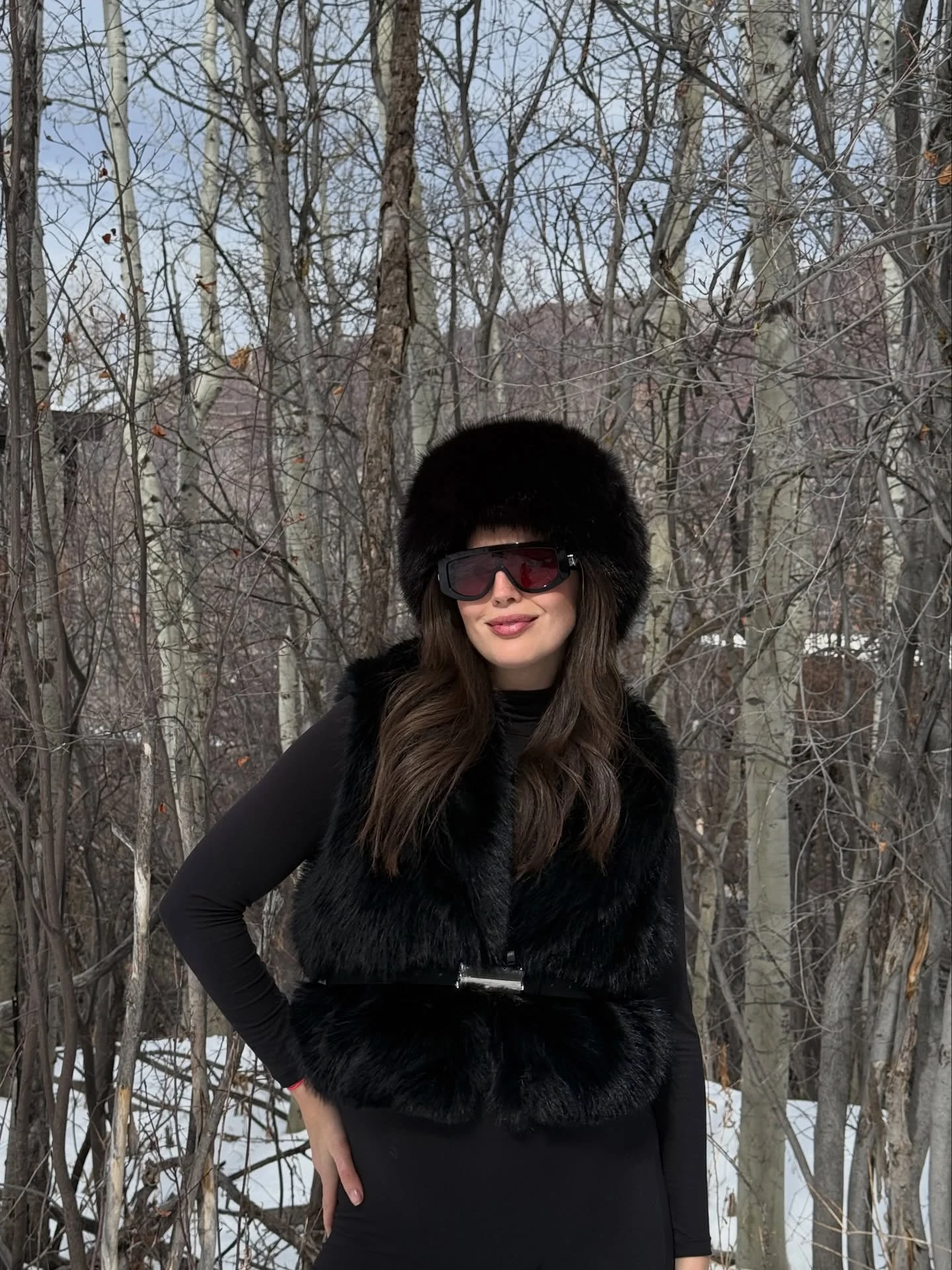 Pregnant woman in black fitted dress, fur vest, fur hat, and dark oversized sunglasses.