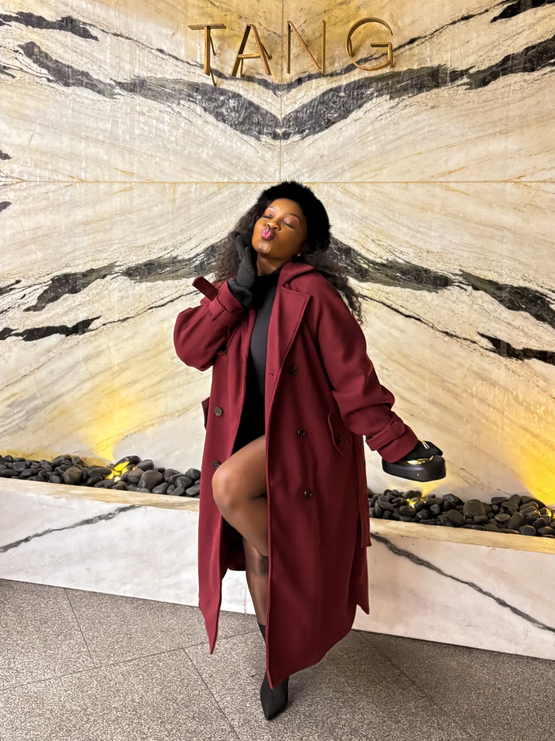 Woman wearing a long burgundy trench coat, black mini dress, fur hat, and black heeled ankle boots.