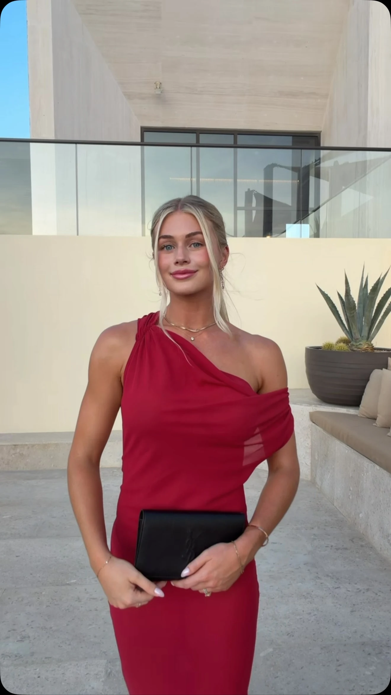 Woman in a fitted cherry red one-shoulder dress holding a black clutch at waist level.