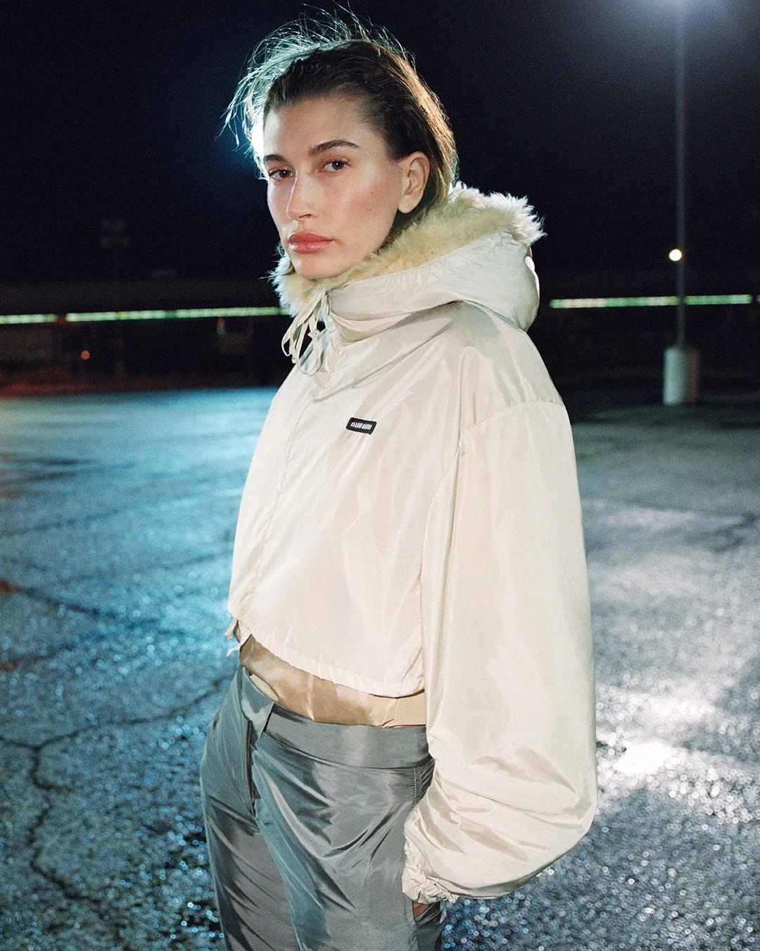 Cream hooded jacket with fluffy trim and shiny silver pants worn outdoors at night.