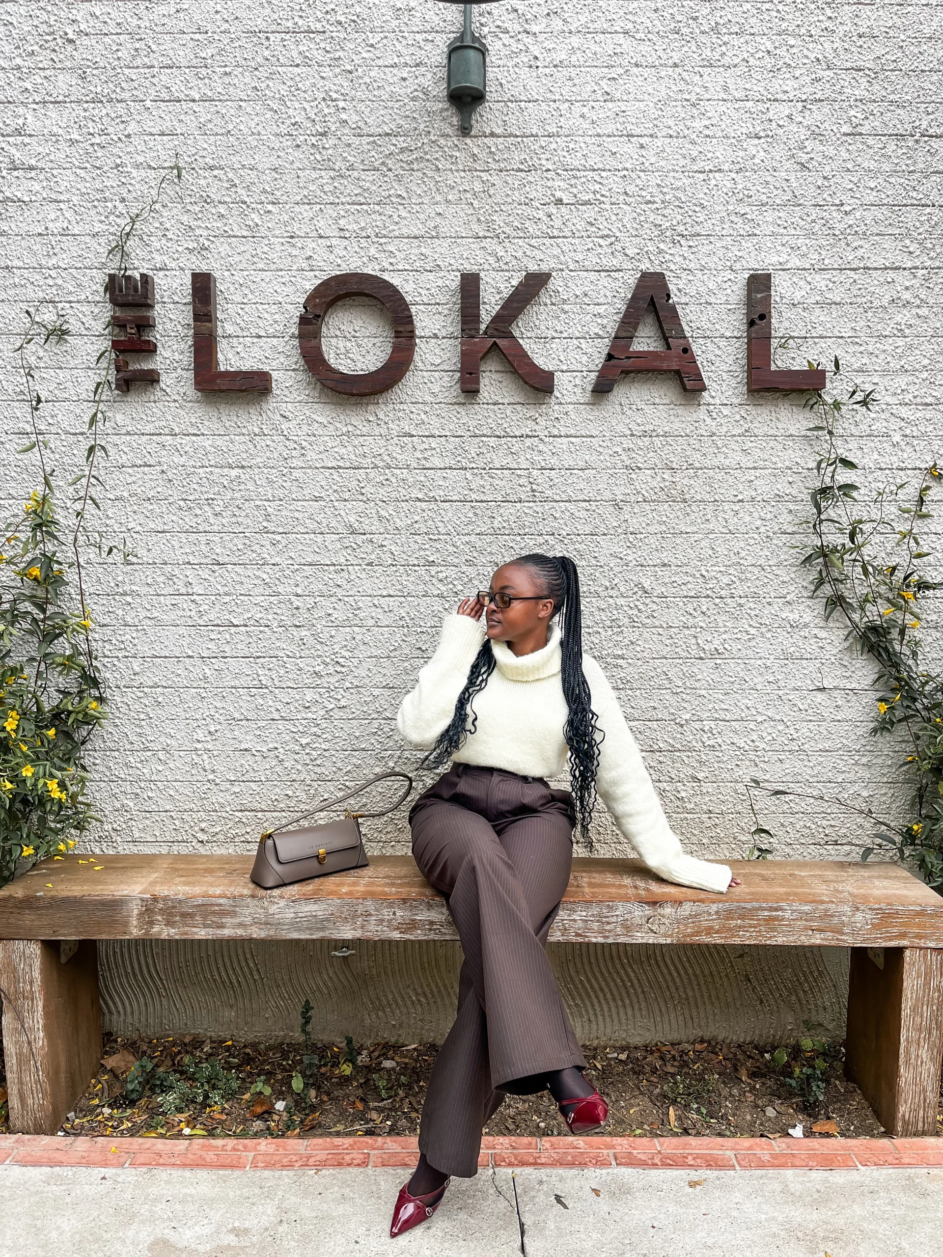 Person in cream turtleneck and dark brown wide-leg trousers with burgundy pointed shoes, seated outdoors.