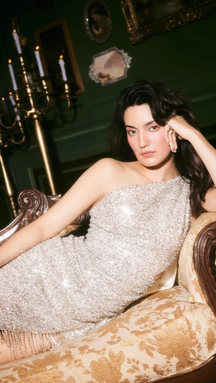 Seated figure in a silver-beige one-shoulder sequined gown with beaded fringe hem.