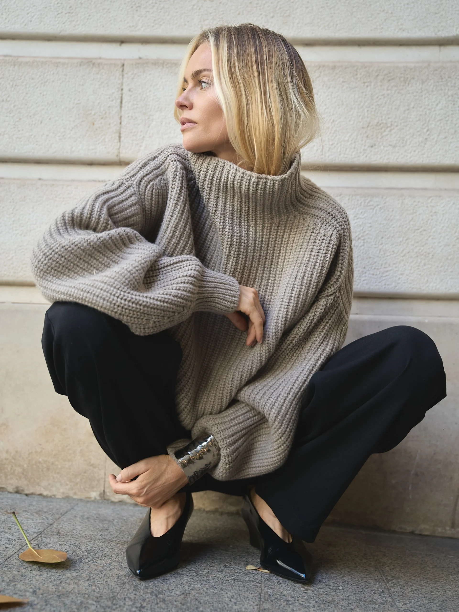 Person crouching by a stone wall in a gray ribbed turtleneck, wide-leg black trousers, and pointed shoes.