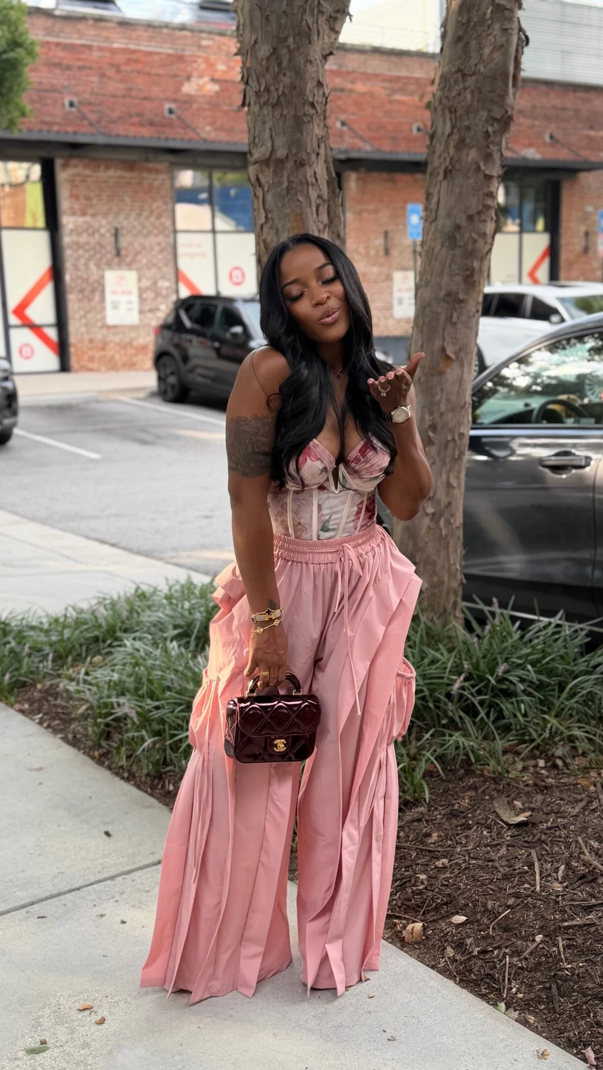 The outfit pairs high-waisted, wide-leg pink trousers with deep vertical pleats and a matching draped sash at the side. A fitted pink top has a ruffled neckline and narrow straps. The trousers fall nearly to the ground and are worn with dark open-toe sandals. Accessories include a small dark burgundy handbag with a short handle and gold-toned hardware, gold-toned bracelets, and a delicate necklace. The wearer has long dark hair, and a tattoo is visible on one upper arm.
