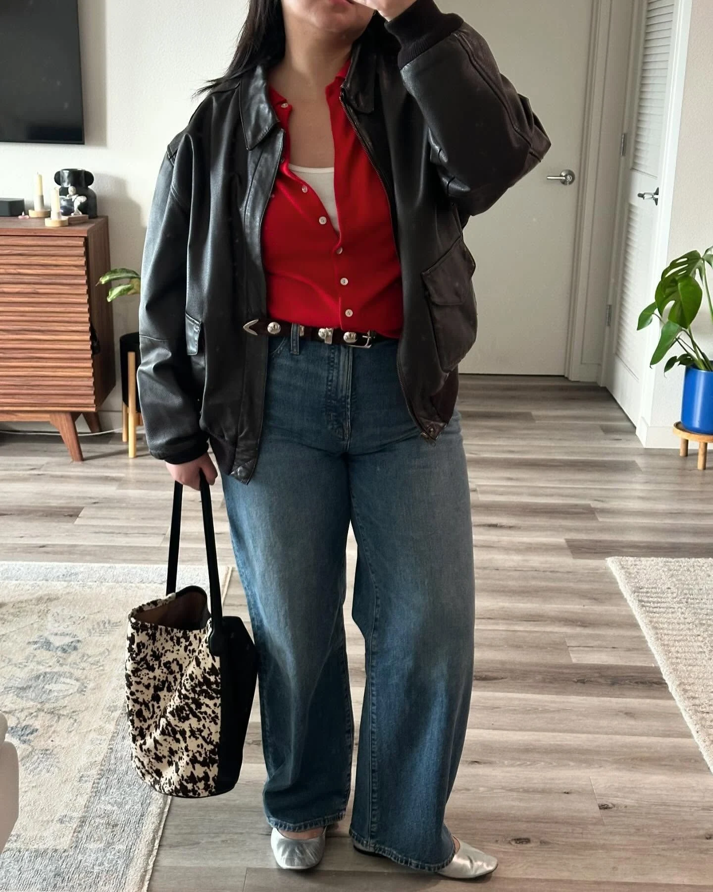 Woman wearing a red cardigan layered under a leather jacket with jeans and a chunky belt.