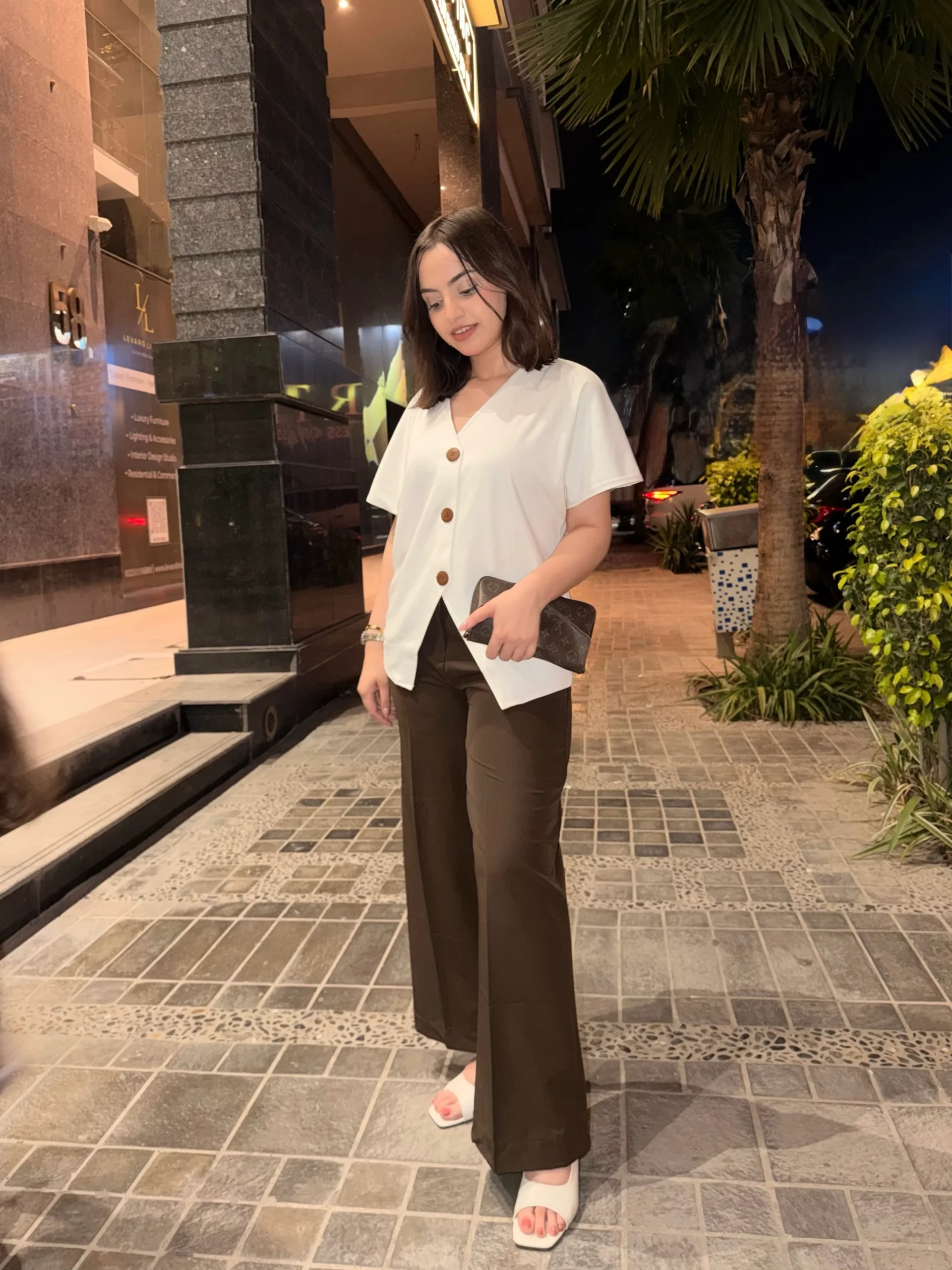 Loose white button blouse with uneven hem paired with dark wide-leg trousers and white sandals.