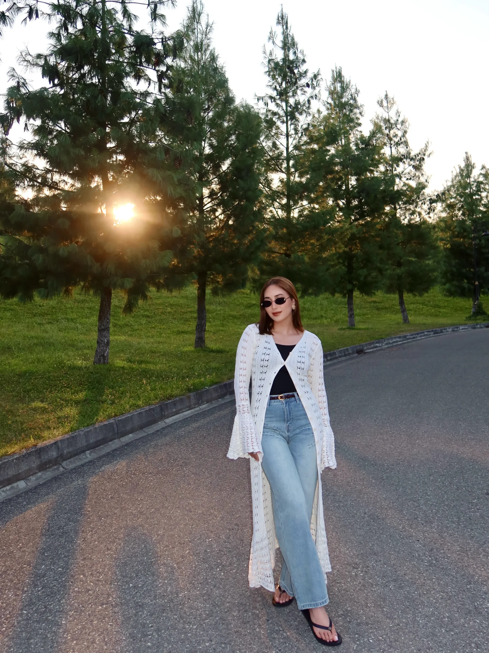 White openwork crochet cardigan over black top, wide-leg jeans, dark belt, and flip-flop sandals.