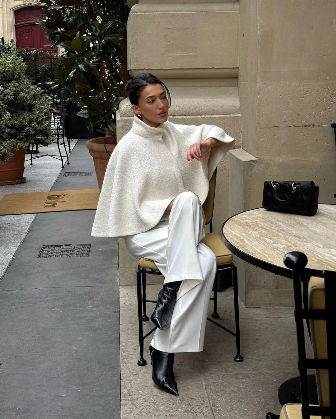 Woman wearing a white textured cape, white tailored pants, and black heeled ankle boots.
