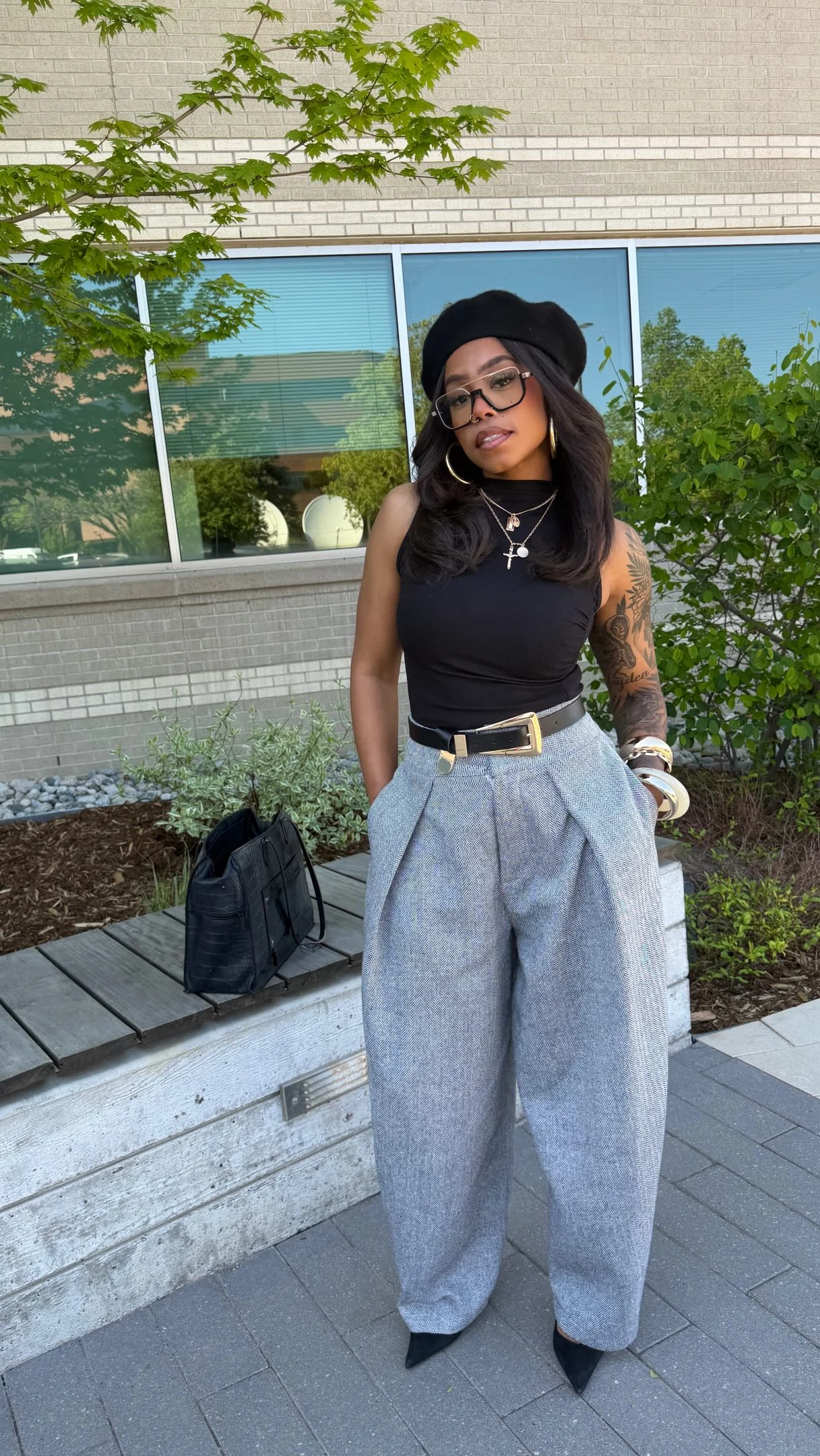 The outfit features a fitted black sleeveless top tucked into high-waisted gray trousers with deep front pleats and very wide, full-length legs. A black belt with a gold-tone buckle defines the waist. The person wears a black brimmed hat, dark rectangular glasses, layered necklaces, and light-colored bracelets. Long dark hair falls over the shoulders, and dark pointed shoes are visible beneath the trouser hems. The trousers have a subtle mottled appearance.