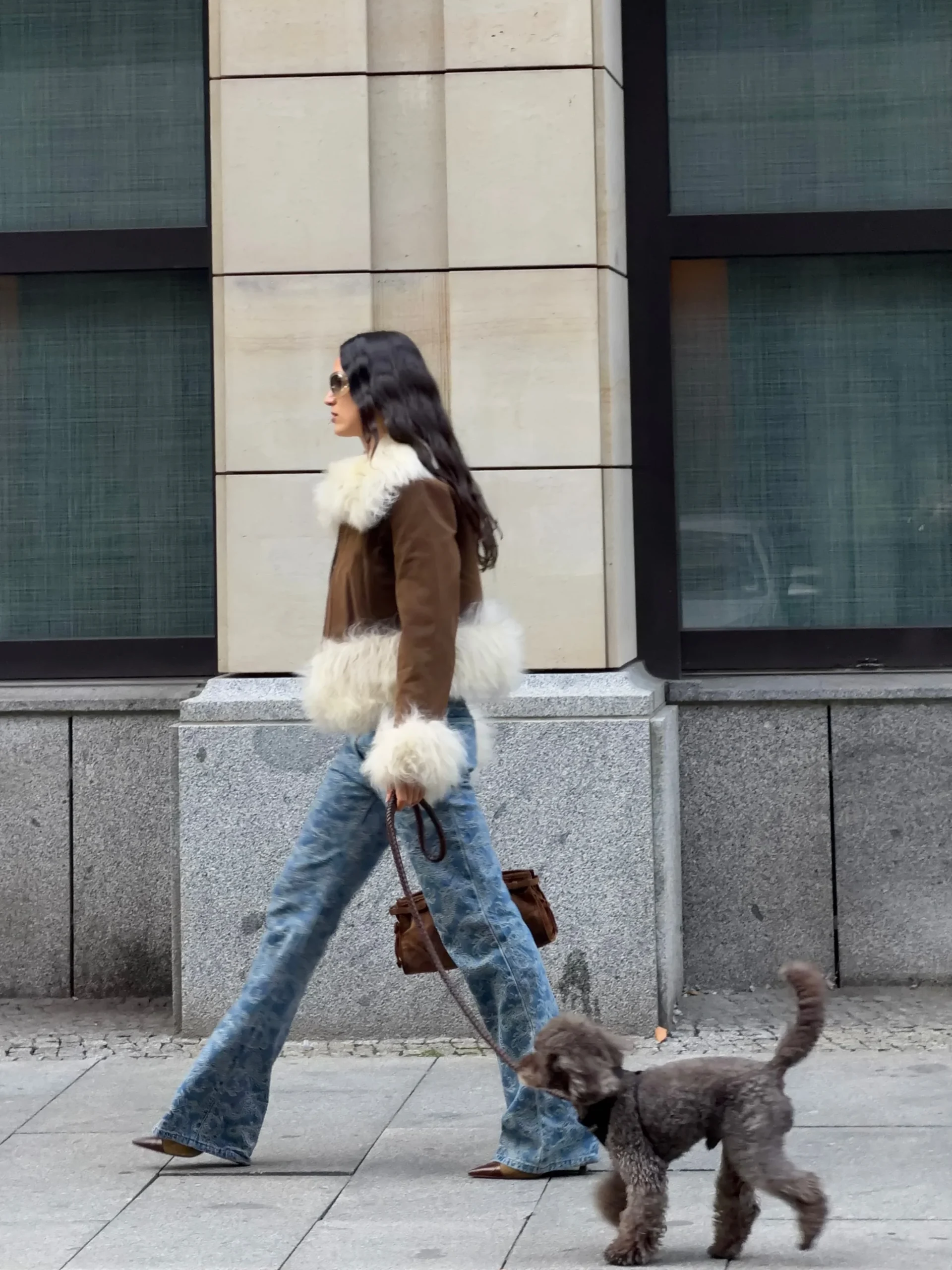 Brown jacket with white fluffy trim worn with blue flared jeans and a small brown handbag.