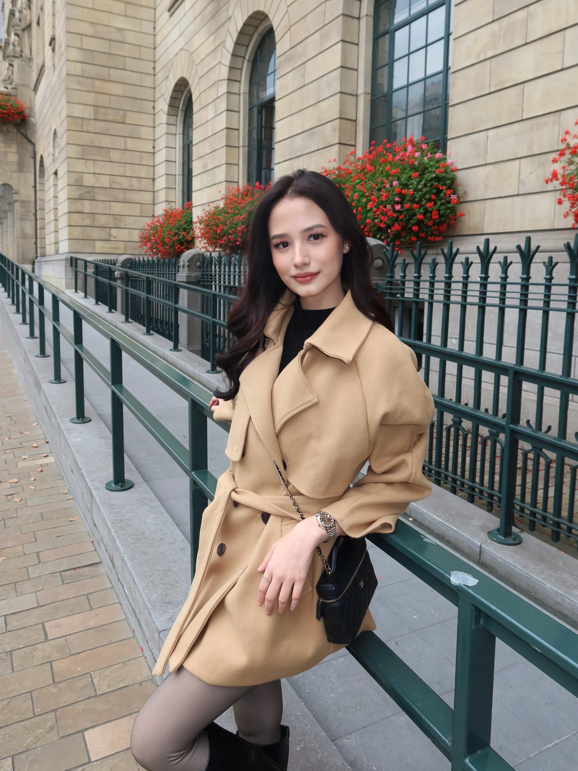 Woman in belted camel coat, black high-neck top, sheer tights, and black ankle boots outdoors.