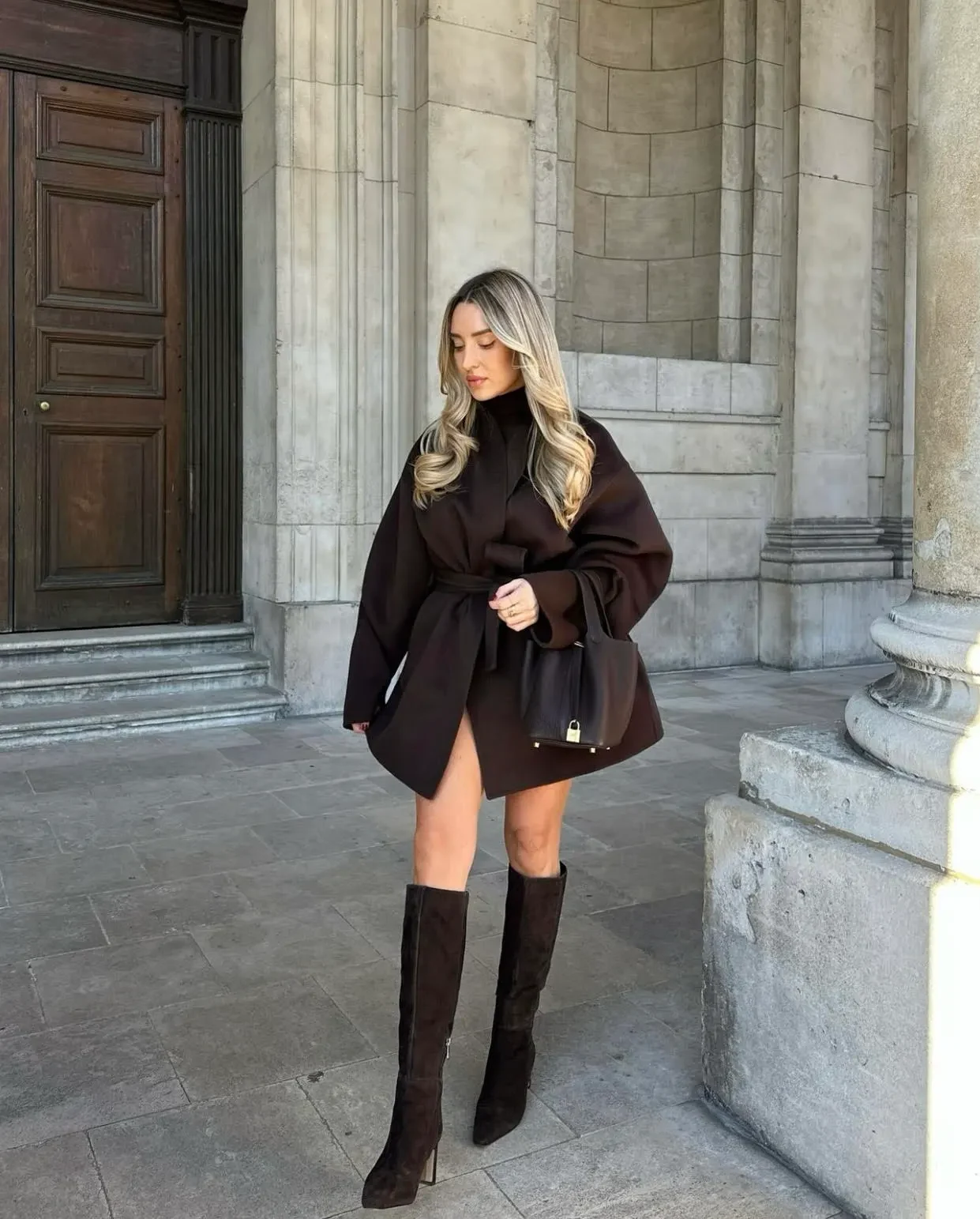 This outfit showcases a rich chocolate-brown wrap coat cinched at the waist for a flattering silhouette. Paired with knee-high suede boots and a sleek black bag, it’s a sophisticated choice for a crisp winter day or evening outing.