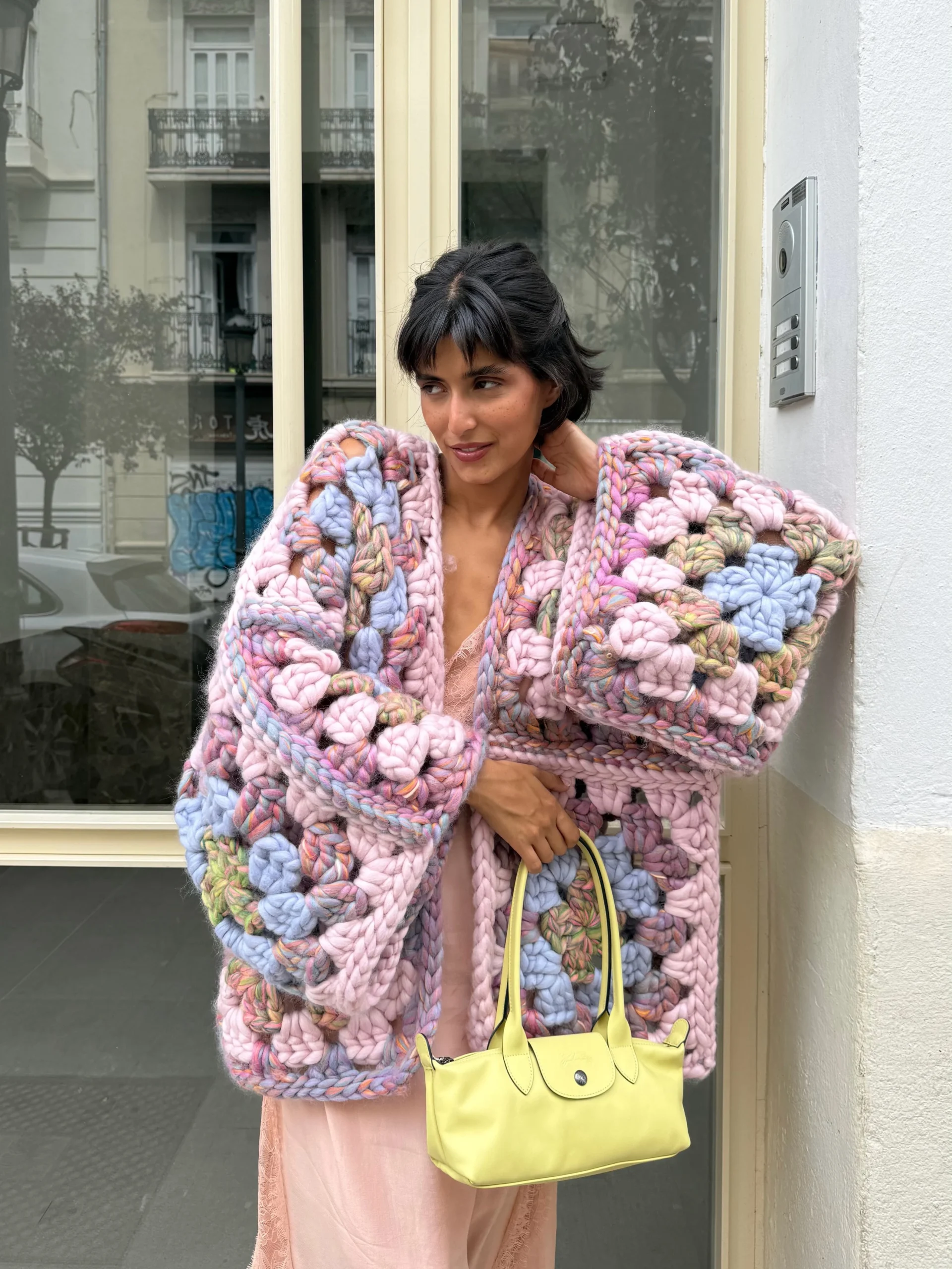 Chunky multicolored crochet cardigan over a pale pink lace dress, carried with a yellow handbag.