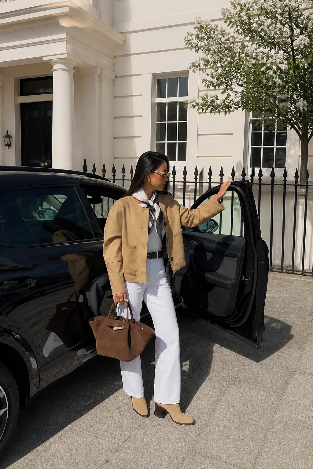 Woman in a cropped tan jacket, white jeans, beige ankle boots, and a brown suede tote.