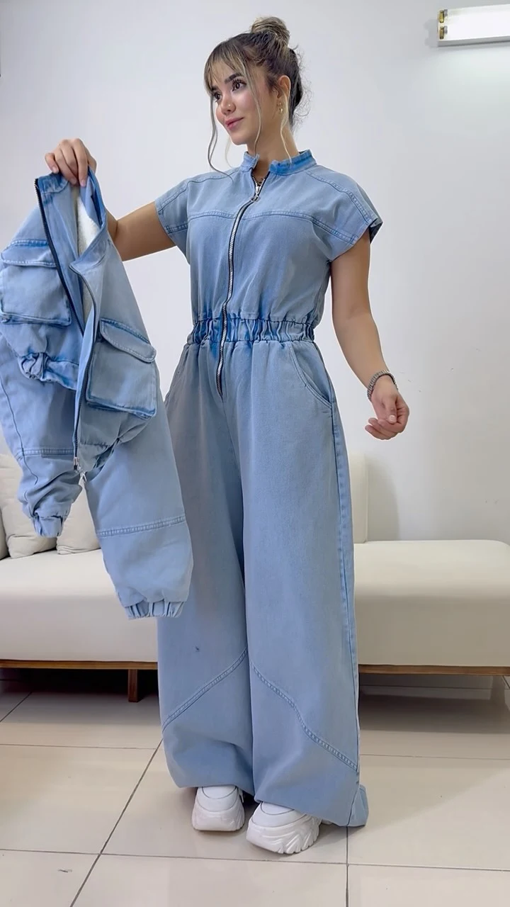 Light blue zip jumpsuit with wide legs and a matching jacket held at the side, white sneakers.