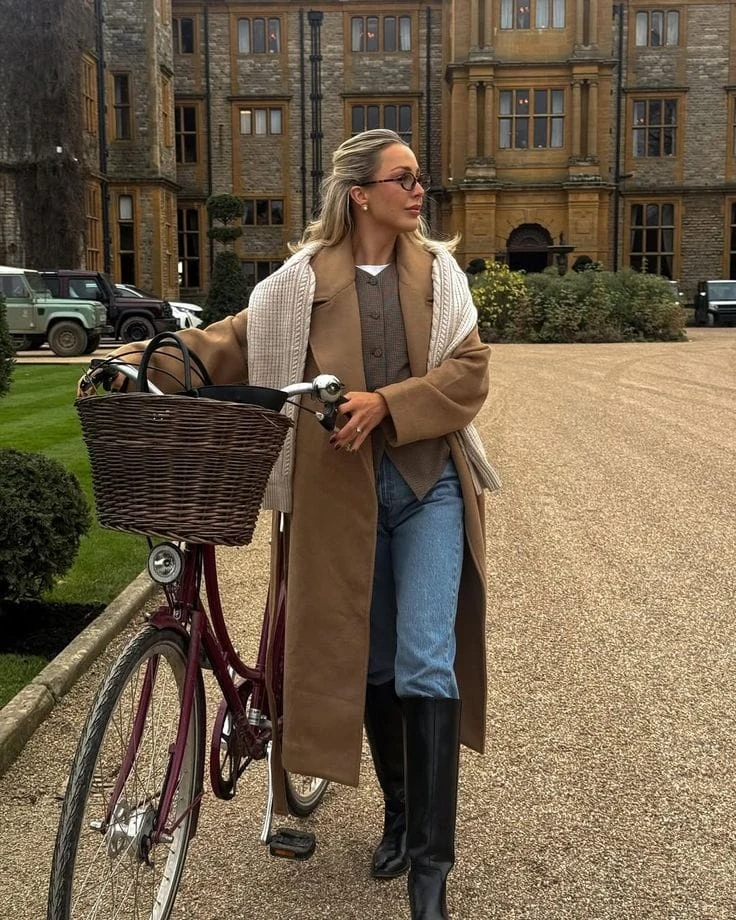 Long camel coat over a beige vest and jeans with a knit scarf and black riding boots.