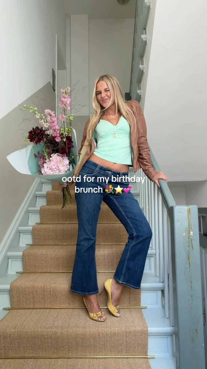 Woman in a mint cropped top, open brown leather jacket, flared jeans, and yellow heeled sandals.
