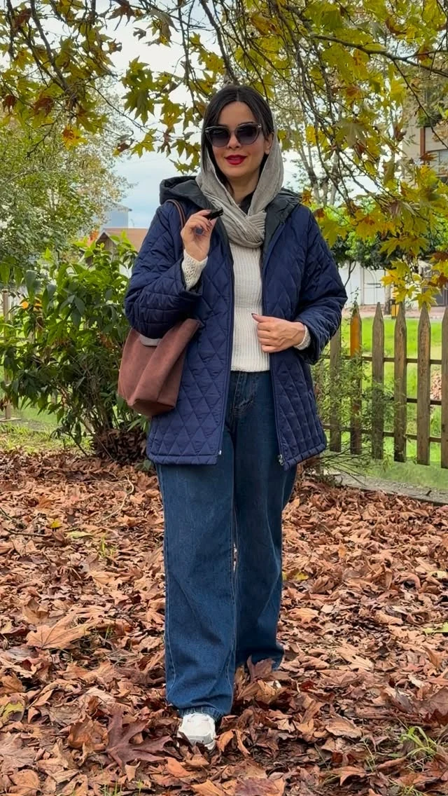 Woman in navy quilted hooded jacket, wide-leg jeans, and white sneakers outdoors among fallen leaves.