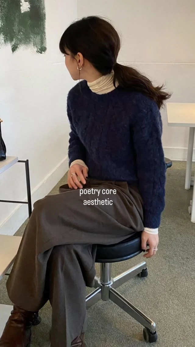 Seated figure wearing a navy sweater over a cream turtleneck with loose brown trousers and dark boots.