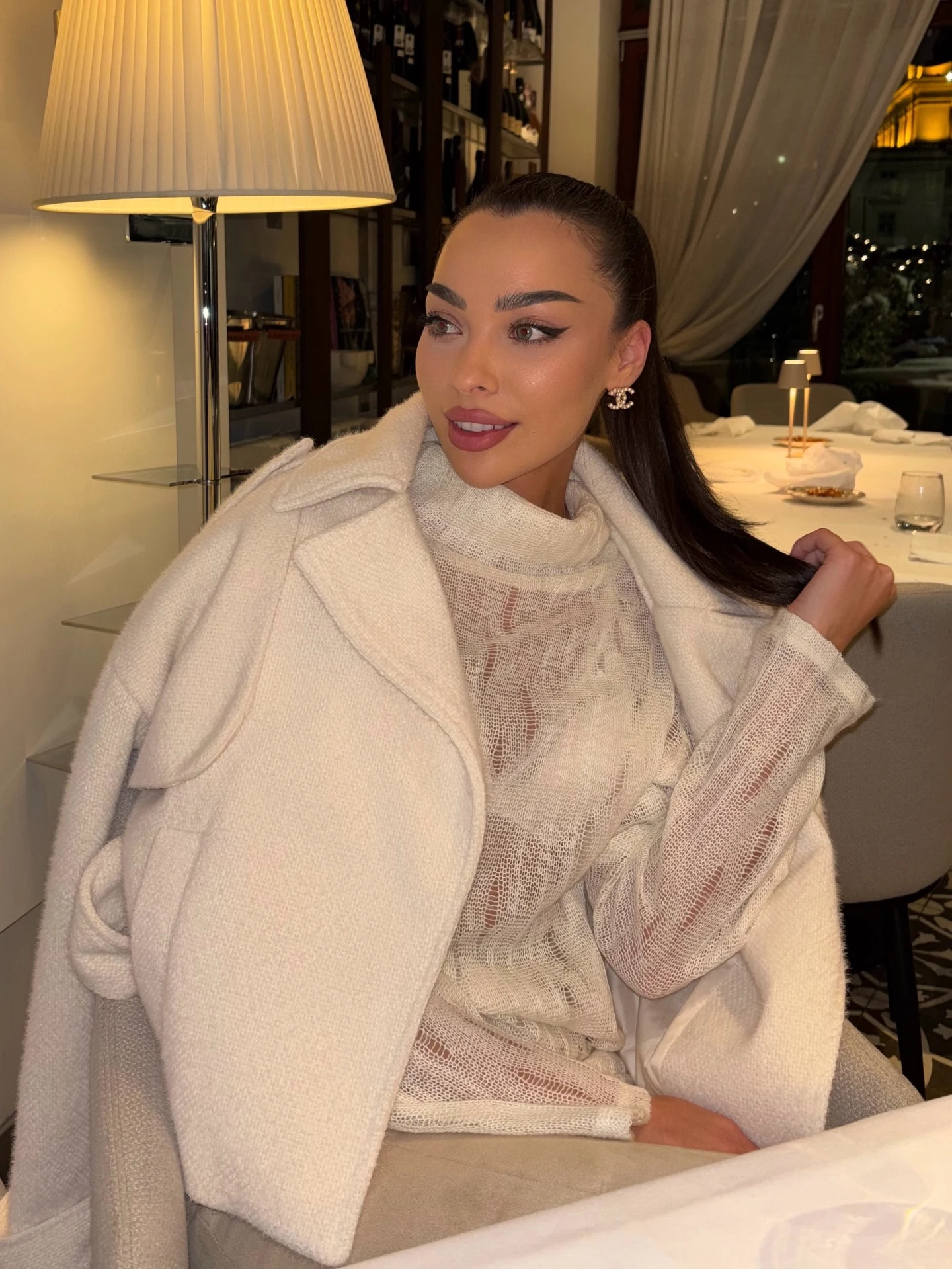 Woman in an oversized cream wool coat layered over a sheer cream knit turtleneck, holding a beige quilted mini bag.