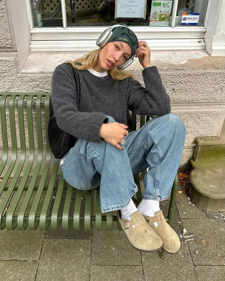 A dark grey oversized knit sweater is paired effortlessly with light-wash baggy cargo jeans for a laid-back, cozy feel. Accessories like a headband, AirPods Max, and suede Birkenstock Boston clogs with white socks add a trendy, casual touch.