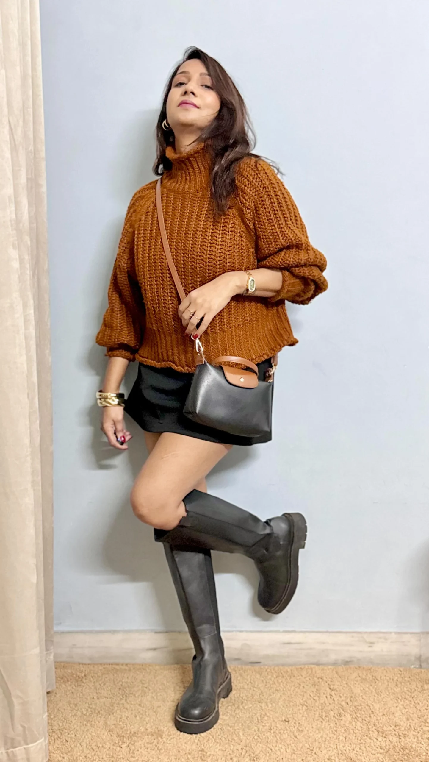 Person wearing a rust knit sweater, short black bottoms, and tall chunky black boots indoors.