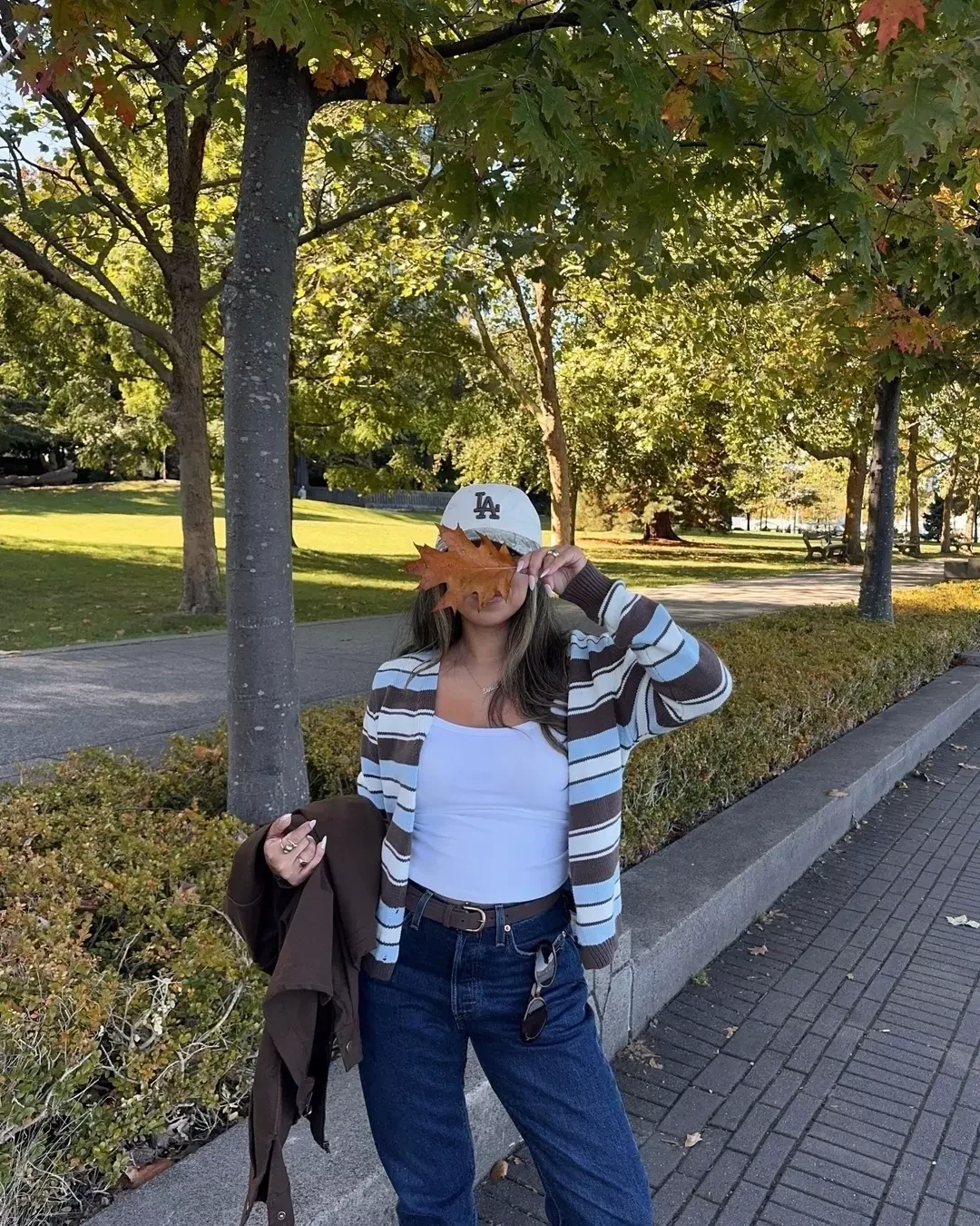 Brown light blue and white striped open cardigan, white ribbed tank top, dark jeans, brown slip on boots, white LA Dodgers cap