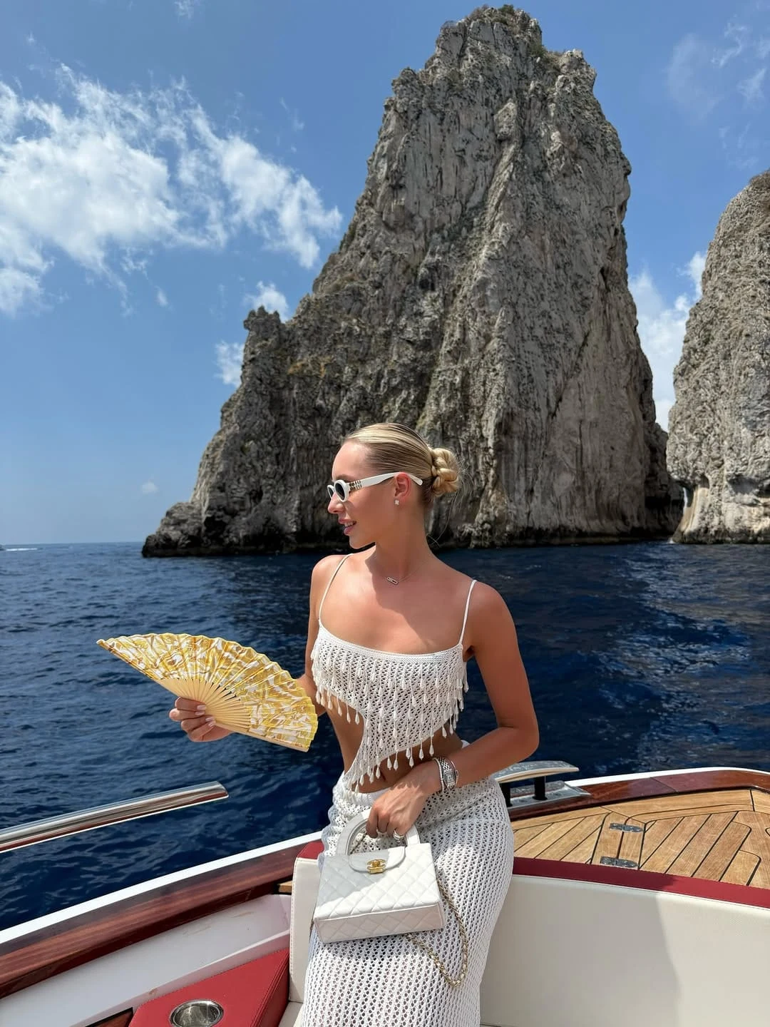 via @anna.bernmark A white crochet crop top with a raw fringe hem is paired with a matching white crochet midi skirt, finished with a white quilted Chanel top-handle bag, a gold fan, white oval sunglasses, and a silver bracelet stack. The fringe hem on the top is the detail that separates this from a standard crochet set it adds movement and a tactile quality that makes the all-white combination feel genuinely considered rather than simply beach-ready. I am completely obsessed with this for a boat day that transitions straight into lunch at a cliffside restaurant. Recreate This Look