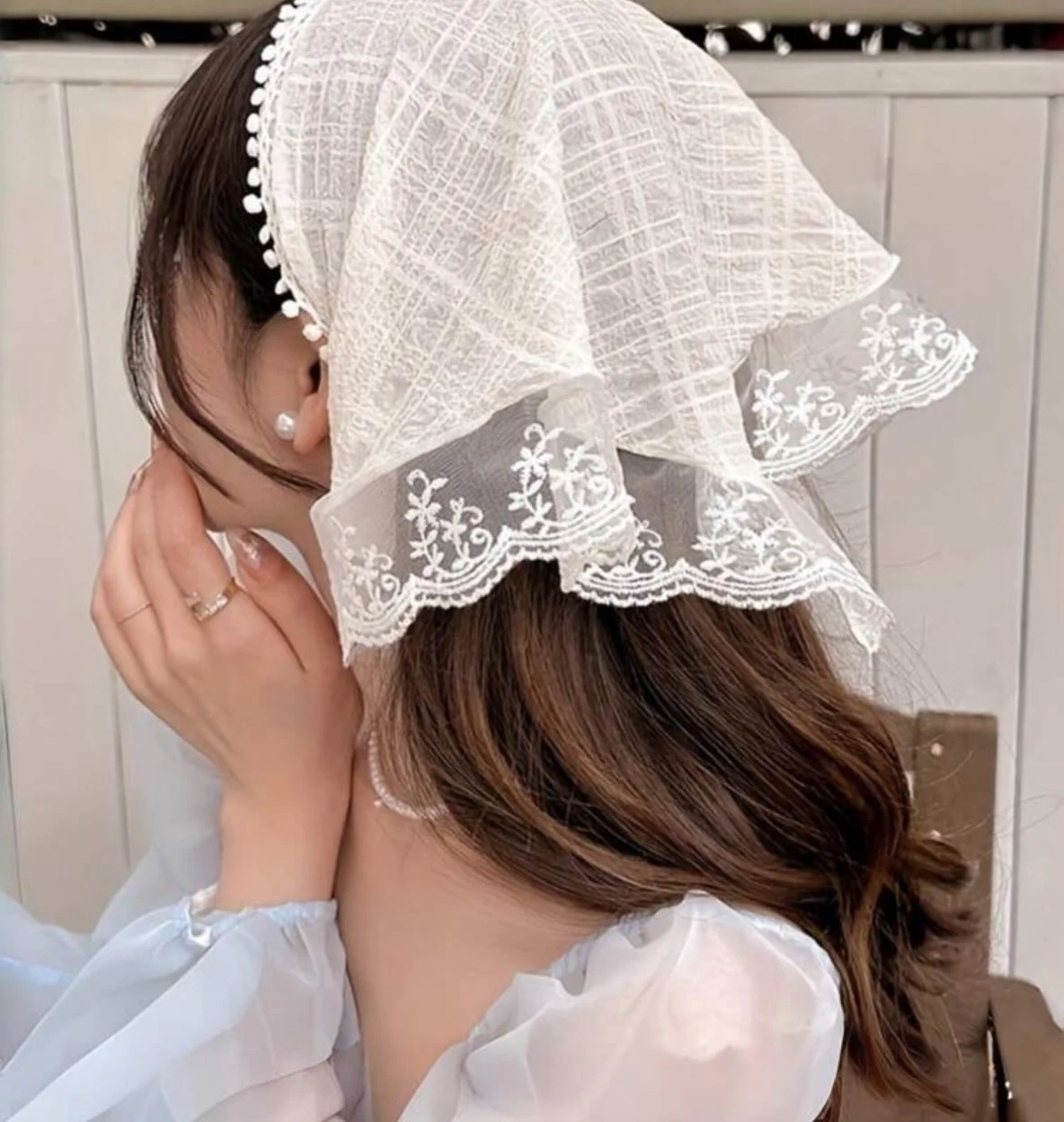 Woman in a white embroidered lace headscarf with pom-pom trim and a pale sheer voluminous blouse.