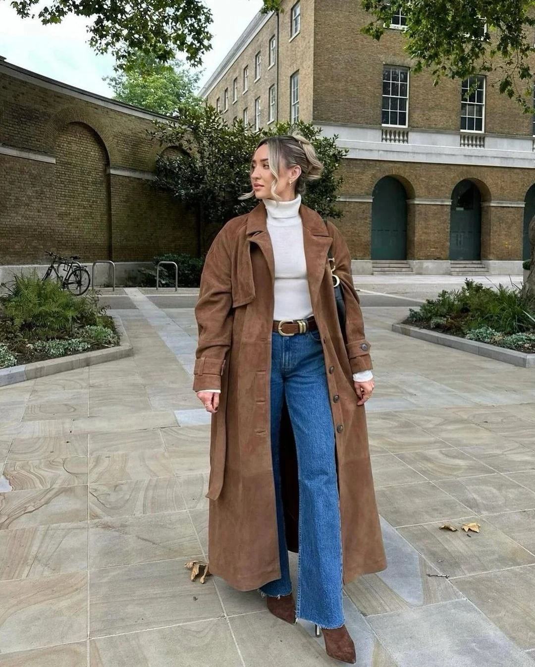 This classic cold-weather ensemble features a brown suede trench coat layered over a white turtleneck, complemented by flared blue jeans for a retro-inspired vibe. The outfit is finished with heeled ankle boots and a chic brown belt, making it perfect for exploring the city in style.