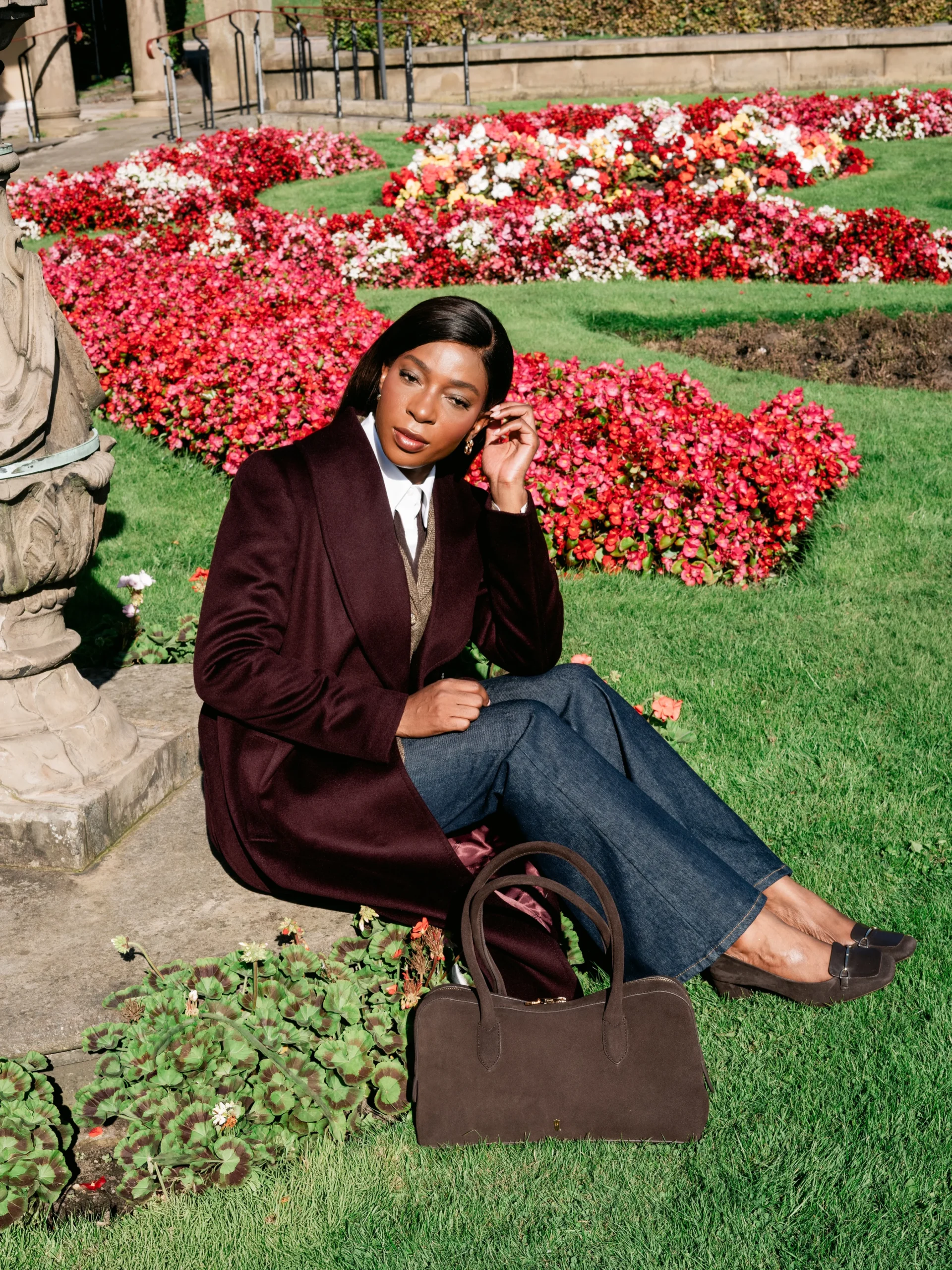 Seated figure wearing a long burgundy coat, white collared shirt, wide-leg jeans, and a dark brown handbag.
